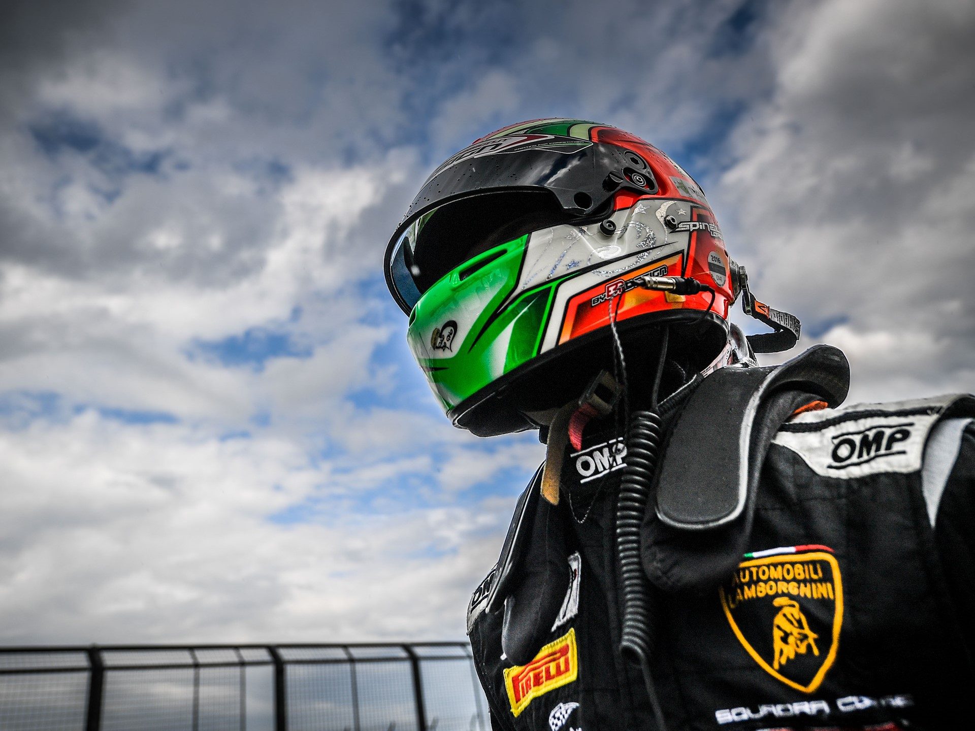 A Lamborghini Squadra Corse racing driver in a multi-colored helmet and black racing suit, photographed from a low angle against a cloudy sky