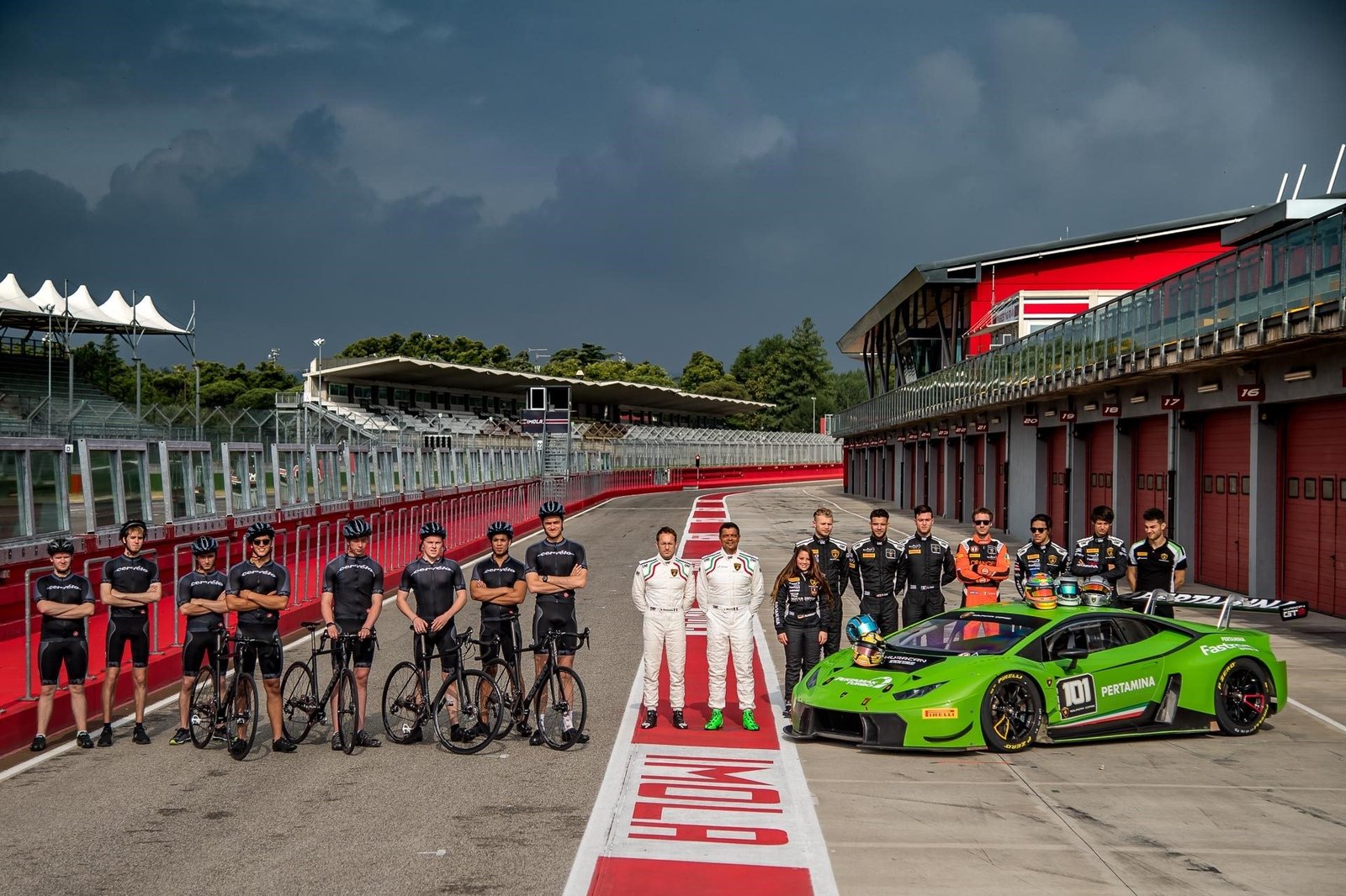 Cyclists and race car drivers pose with a green Lamborghini Huracán GT3 on the Imola Autodrome pit lane under dramatic skies