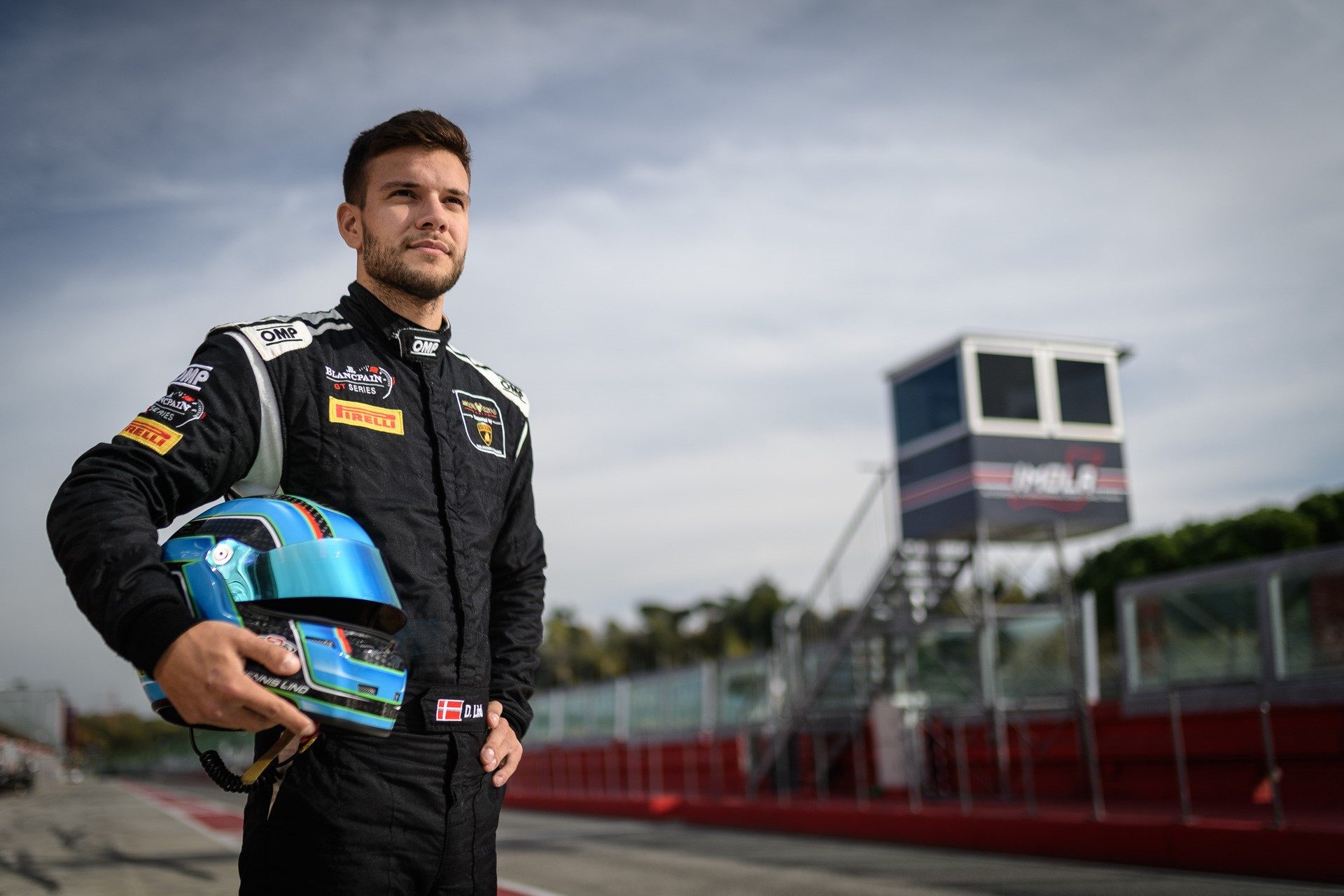 Dennis Lind standing on the Imola racetrack in a black racing suit, holding a blue helmet with the control tower visible behind him