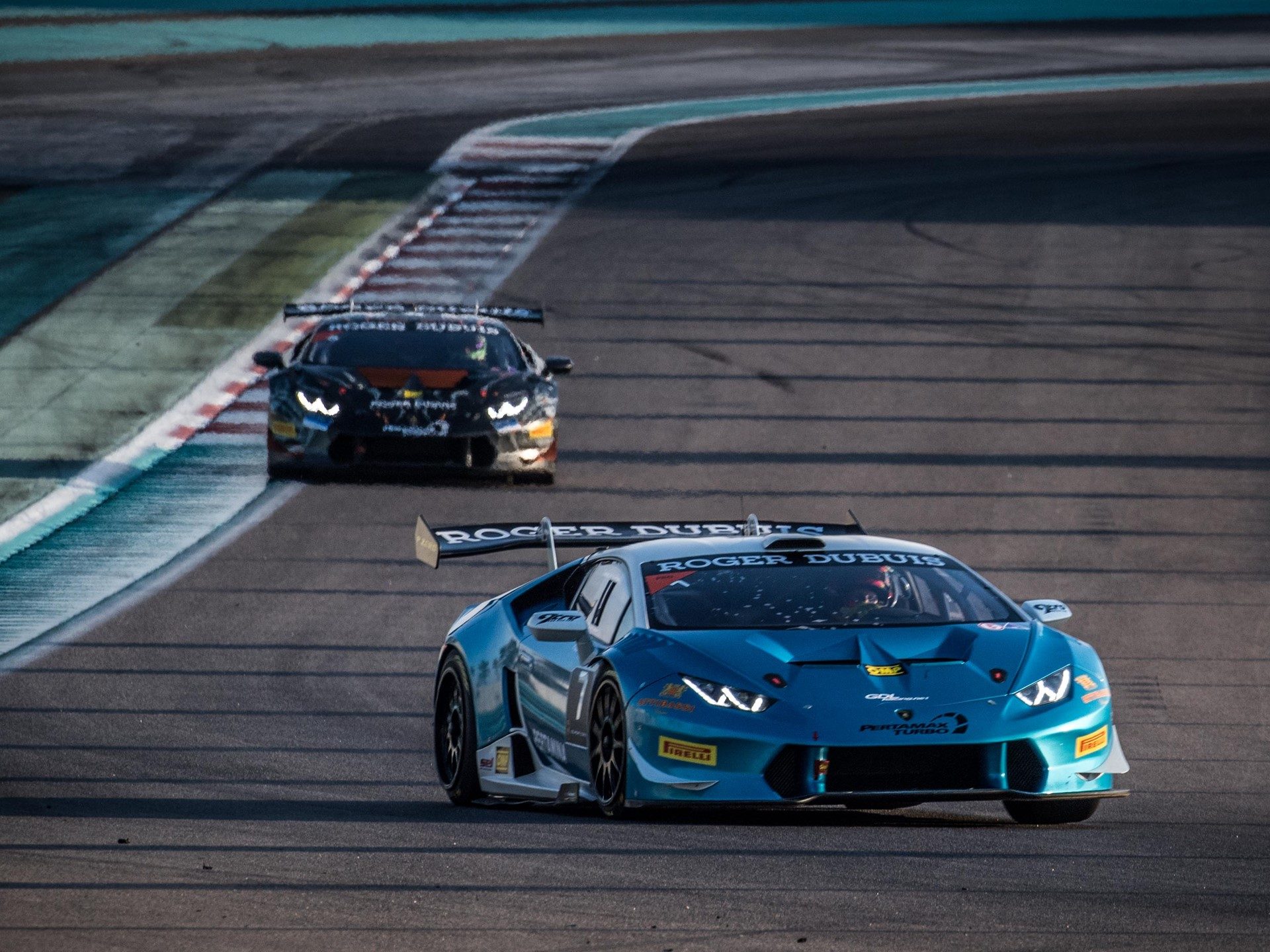Two Lamborghini Huracán Super Trofeo race cars battling for position on track, blue car leading a black car, both featuring full racing liveries and large rear wings