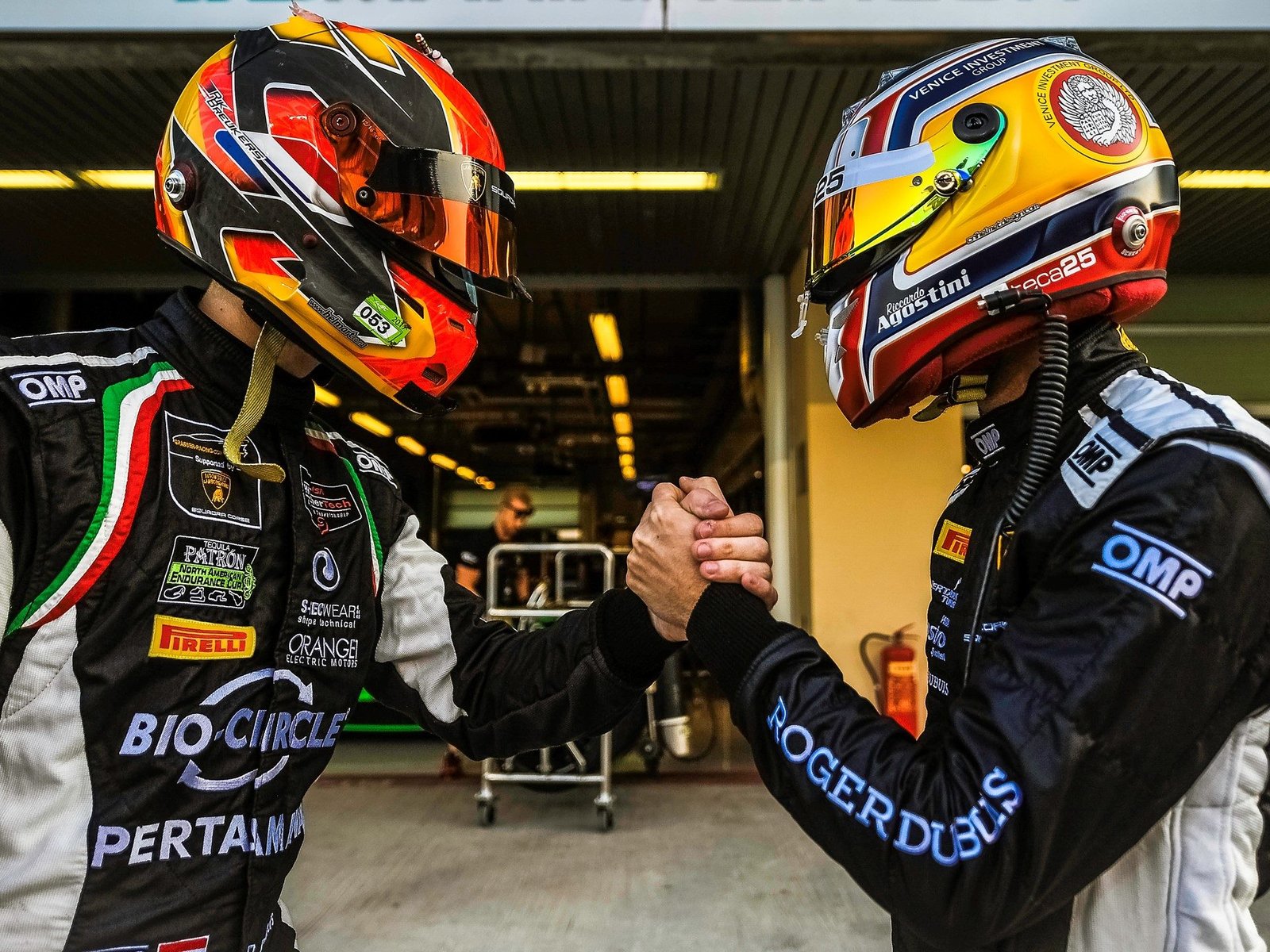 Two Lamborghini Squadra Corse drivers in racing suits and helmets shaking hands in the pit lane, with Squadra Corse branding visible on their gear