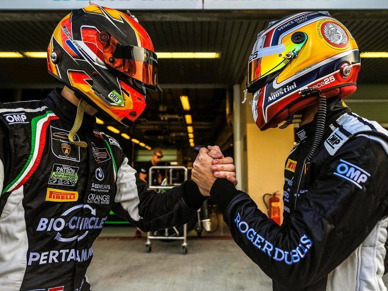Two Lamborghini Squadra Corse drivers in racing suits and helmets shaking hands in the pit lane, with Squadra Corse branding visible on their gear