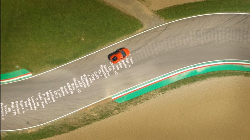 Aerial view of an orange Lamborghini driving on a racetrack with YouTube subscriber names projected onto the asphalt