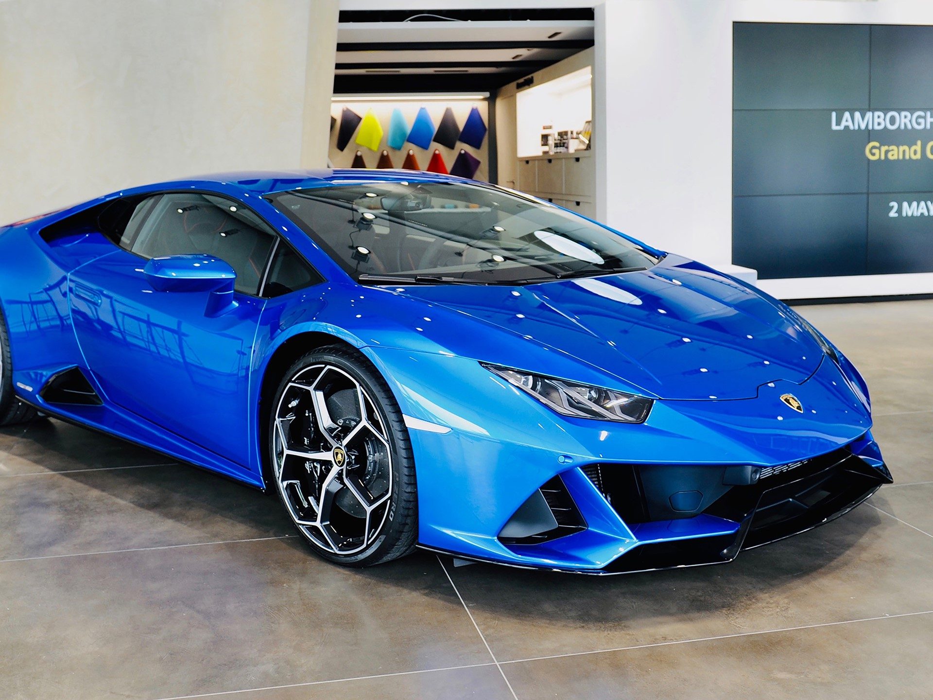 A vibrant blue Lamborghini Huracán displayed in a brightly lit modern showroom with sharp angular design lines and Y-shaped daytime running lights