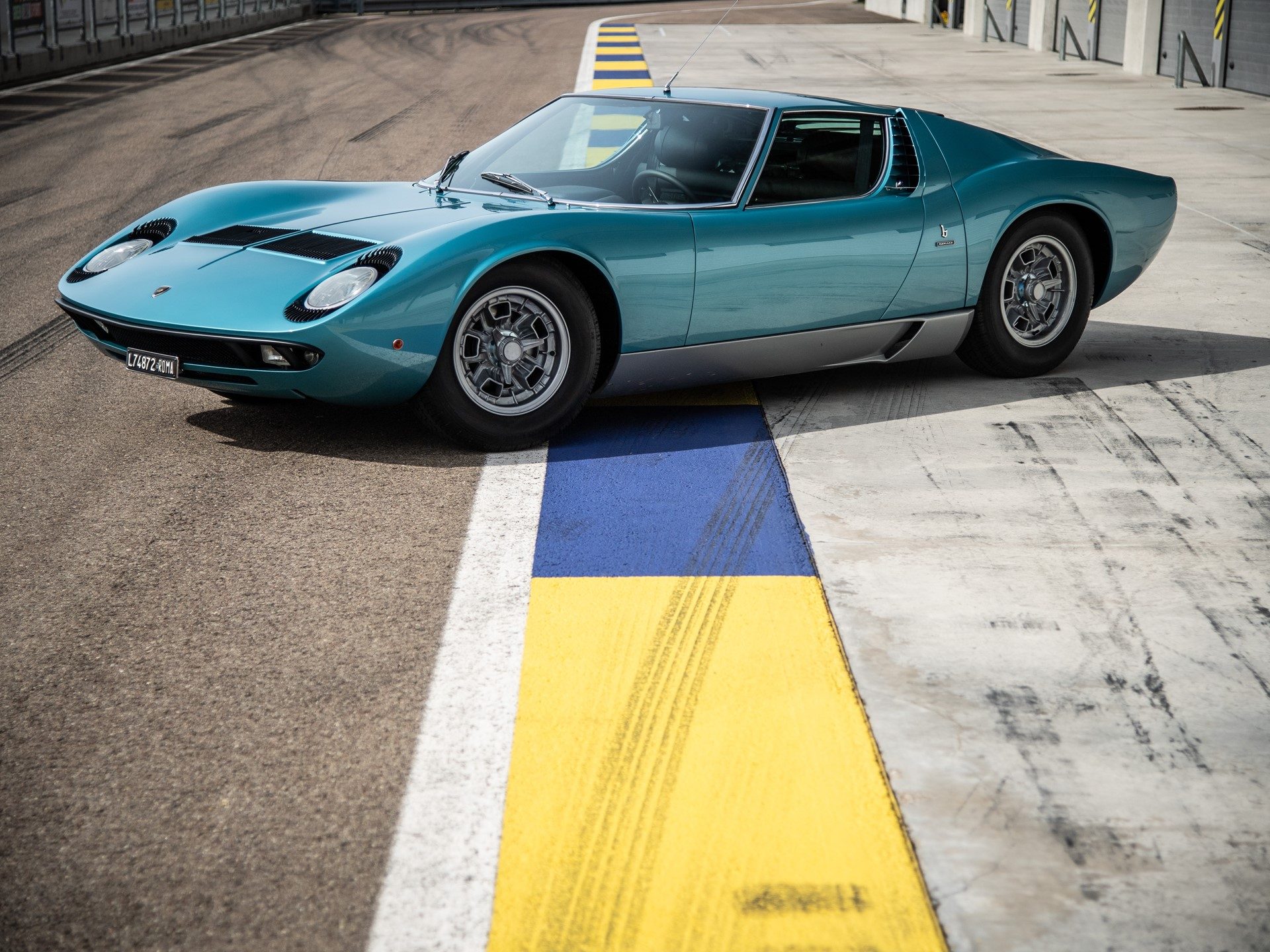 Lamborghini Miura P400 in Azzurro Mexico blue parked on a racetrack, showcasing its iconic mid-engine profile and pop-up headlights