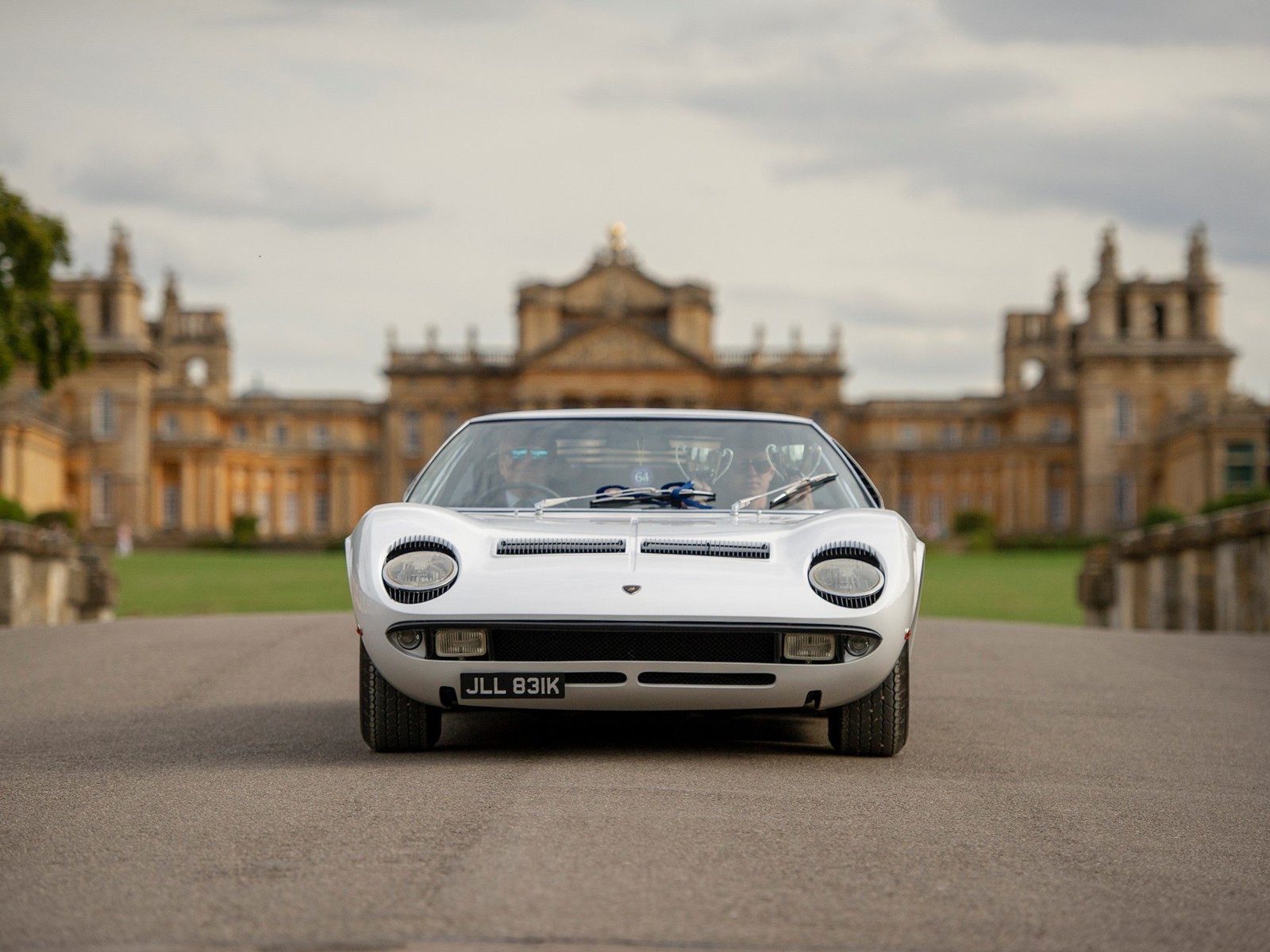 A silver Lamborghini Miura S with a concours trophy visible through the windshield, parked in front of a grand classical building