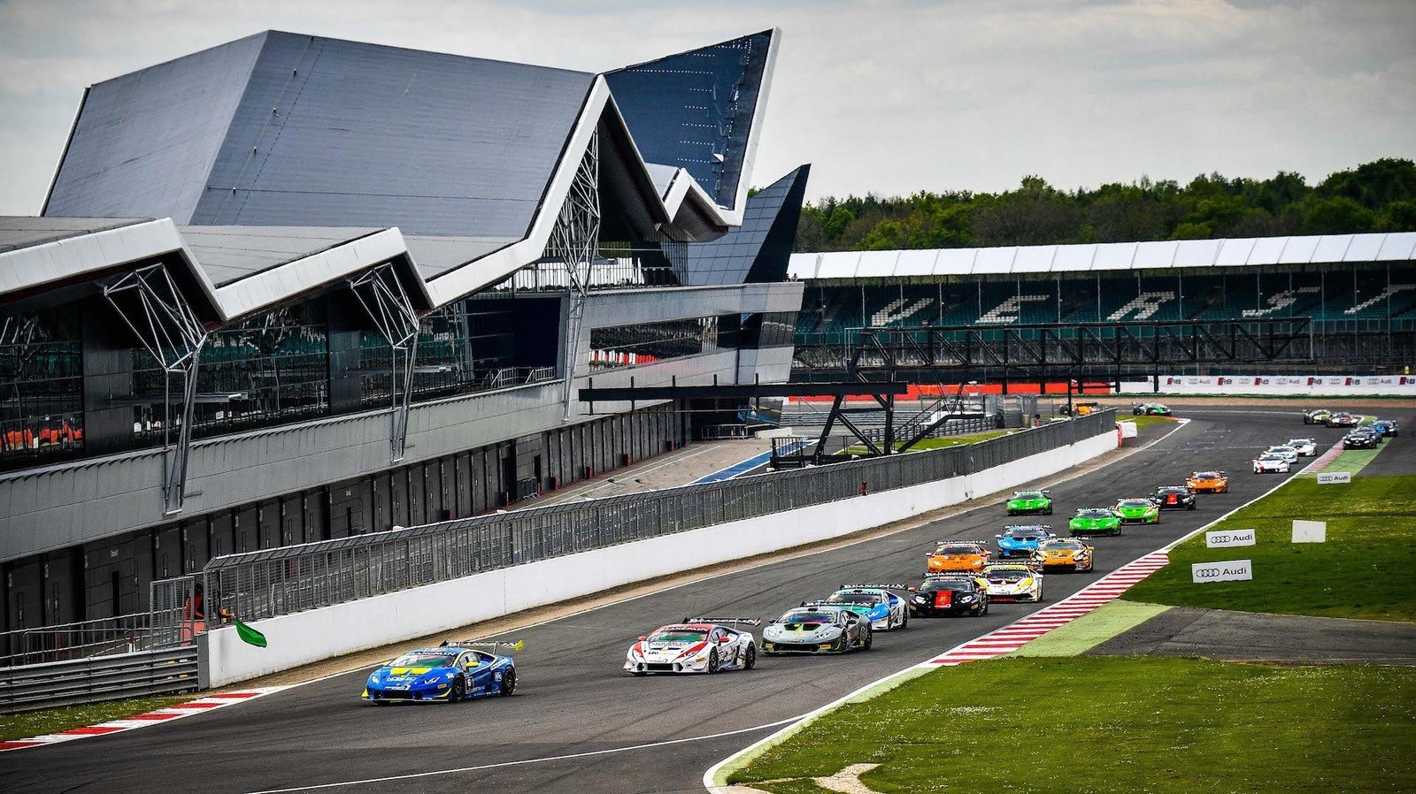A pack of Lamborghini Huracán Super Trofeo race cars in various liveries battling through a corner at Silverstone under overcast skies