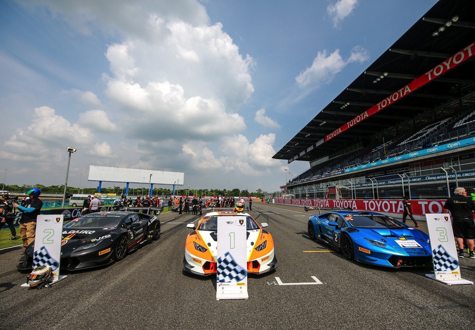 Three Lamborghini Huracán Super Trofeo race cars in black, orange/white, and blue liveries lined up on the grid at Chang International Circuit in Buriram, Thailand
