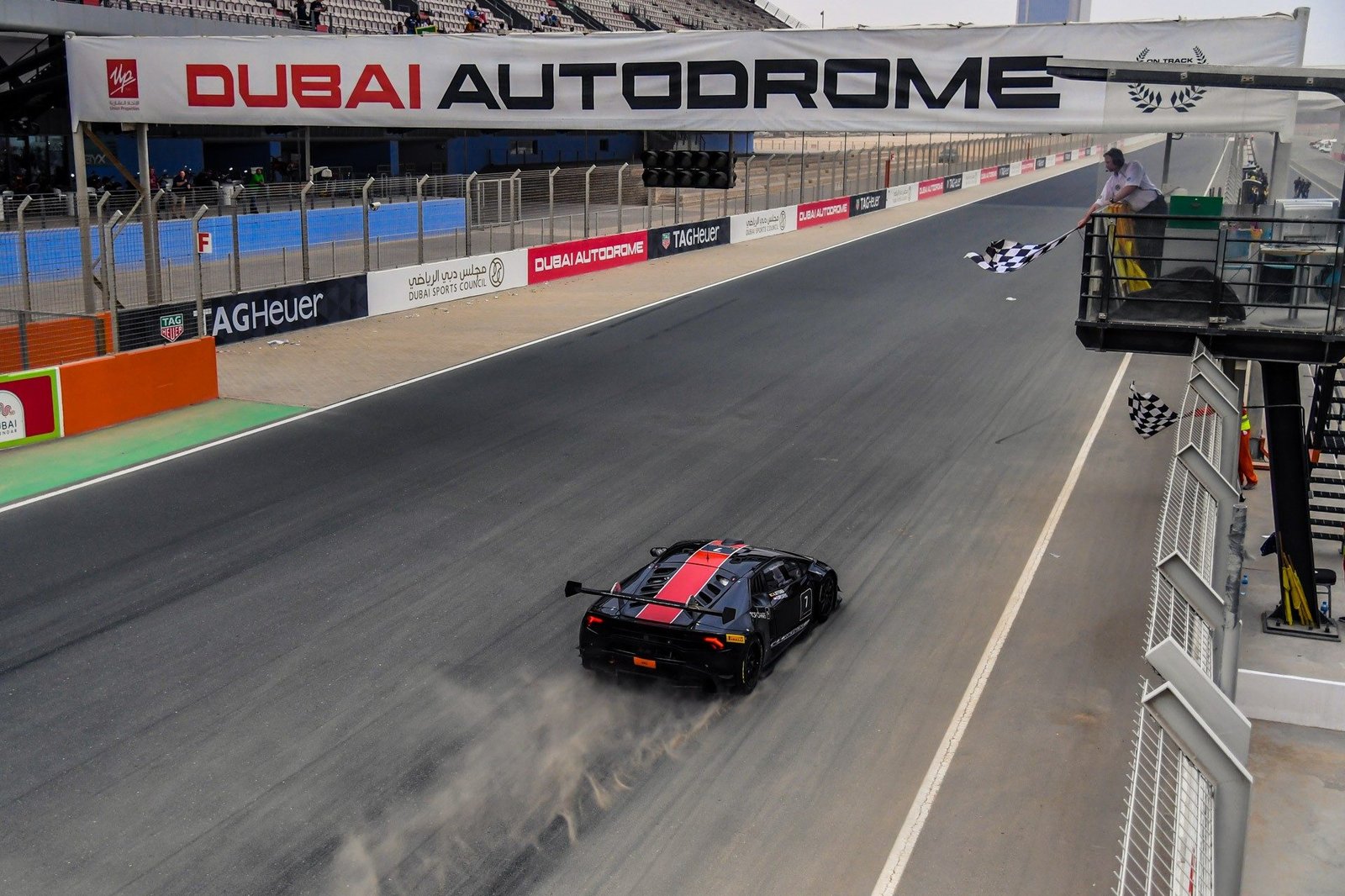 A black Lamborghini Huracán Super Trofeo race car with red stripes crosses the finish line under the checkered flag at Dubai Autodrome