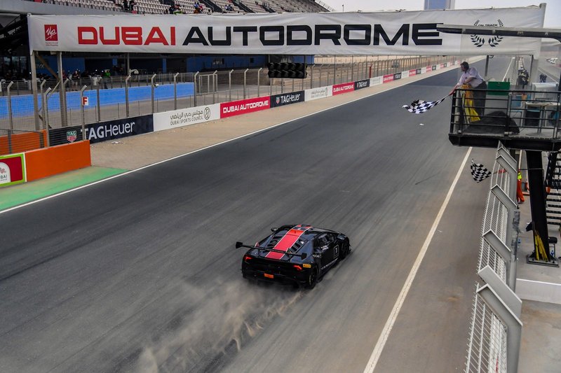 A black Lamborghini Huracán Super Trofeo race car with red stripes crosses the finish line under the checkered flag at Dubai Autodrome