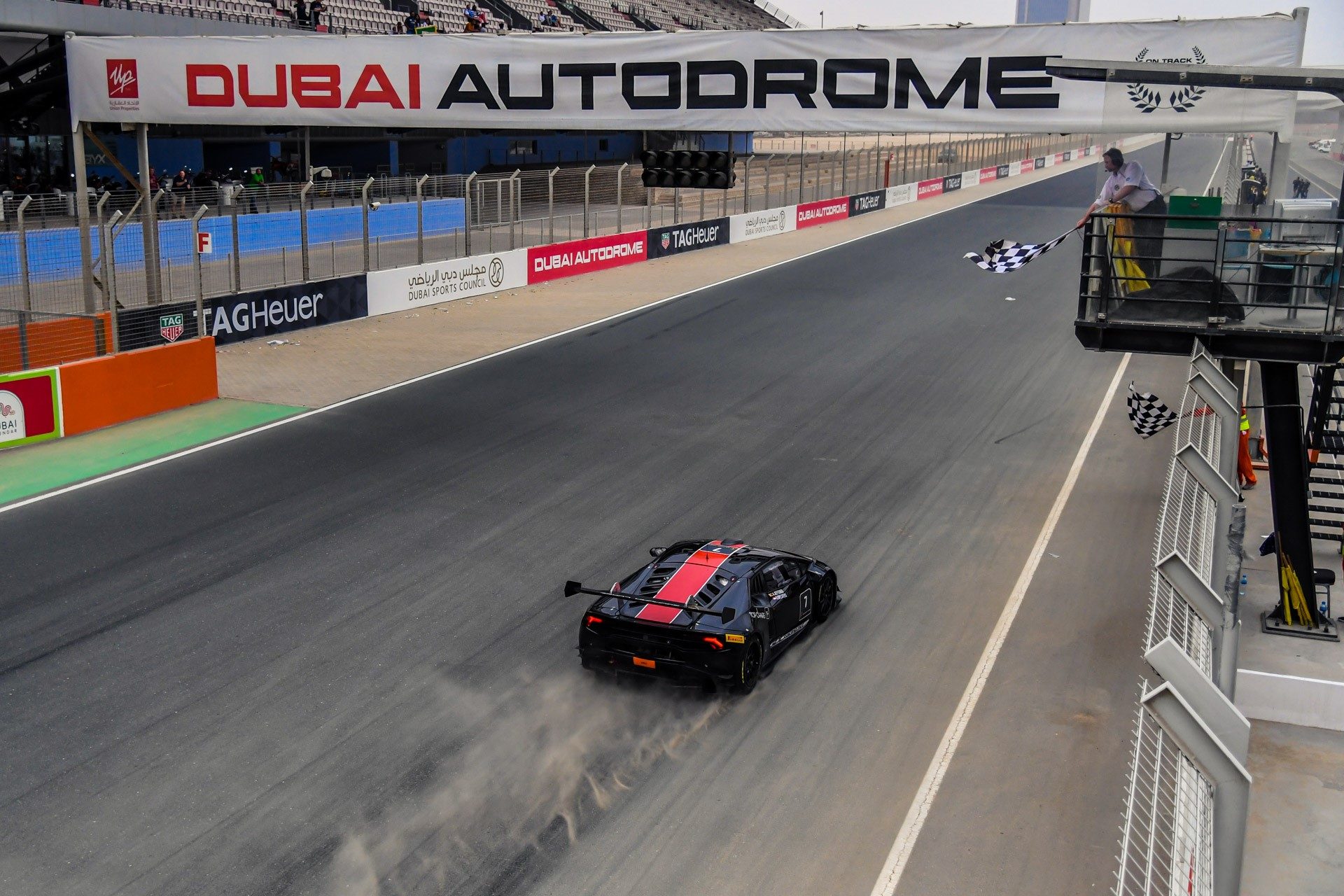 A black Lamborghini Huracán Super Trofeo race car with red stripes crosses the finish line under the checkered flag at Dubai Autodrome