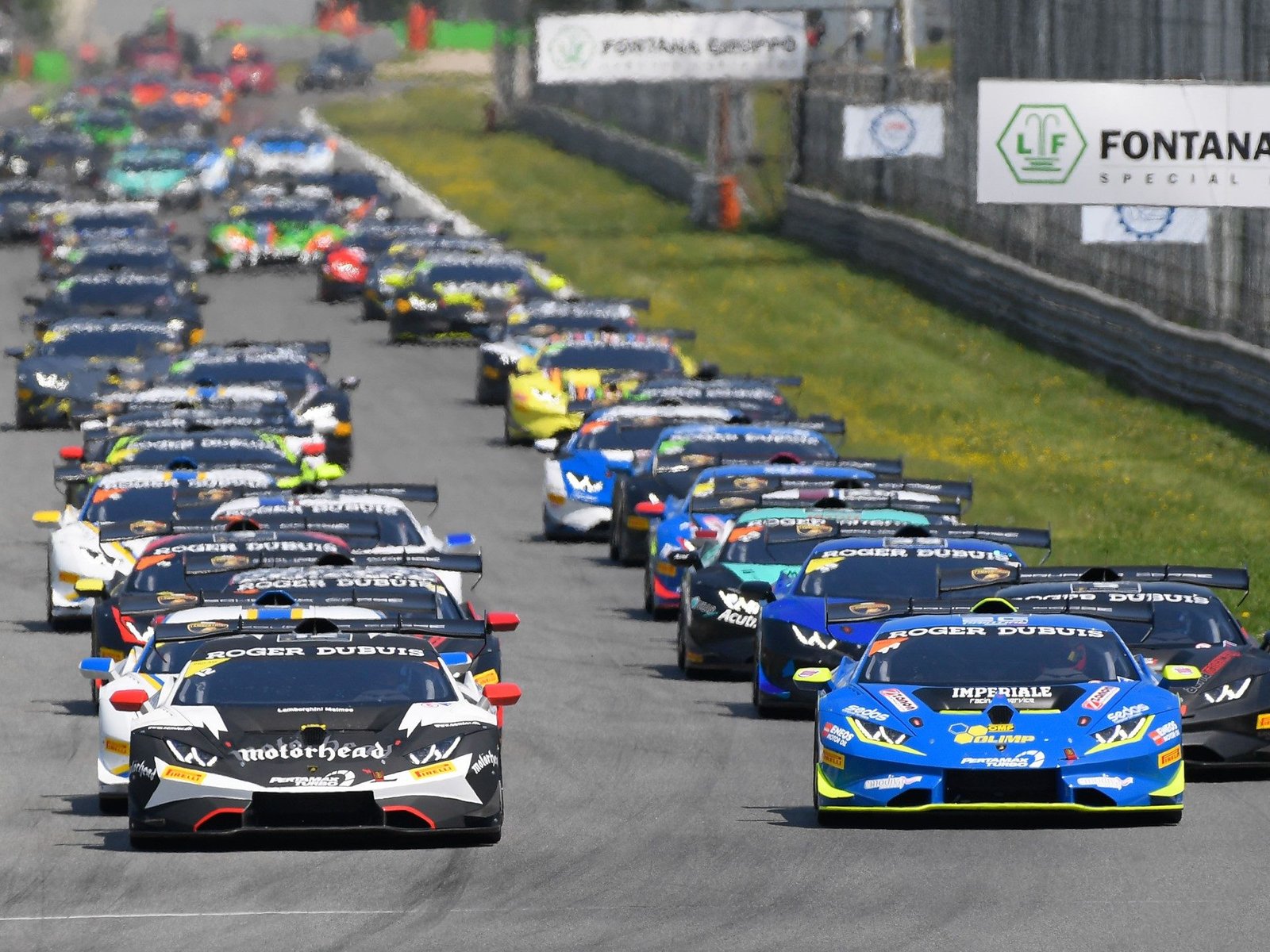 Multiple Lamborghini Huracán Super Trofeo Evo race cars in blue, black, and white liveries racing closely together at Monza with sponsor banners visible trackside