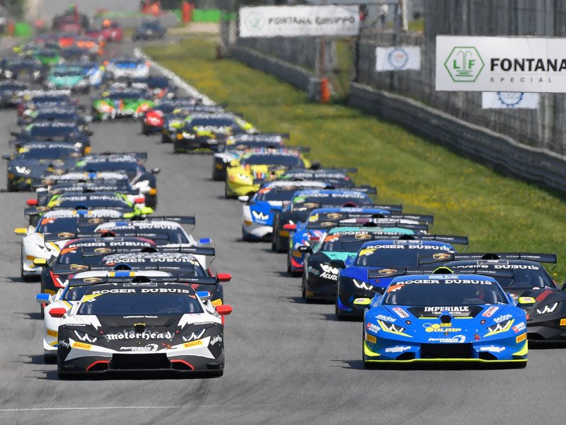 Multiple Lamborghini Huracán Super Trofeo Evo race cars in blue, black, and white liveries racing closely together at Monza with sponsor banners visible trackside