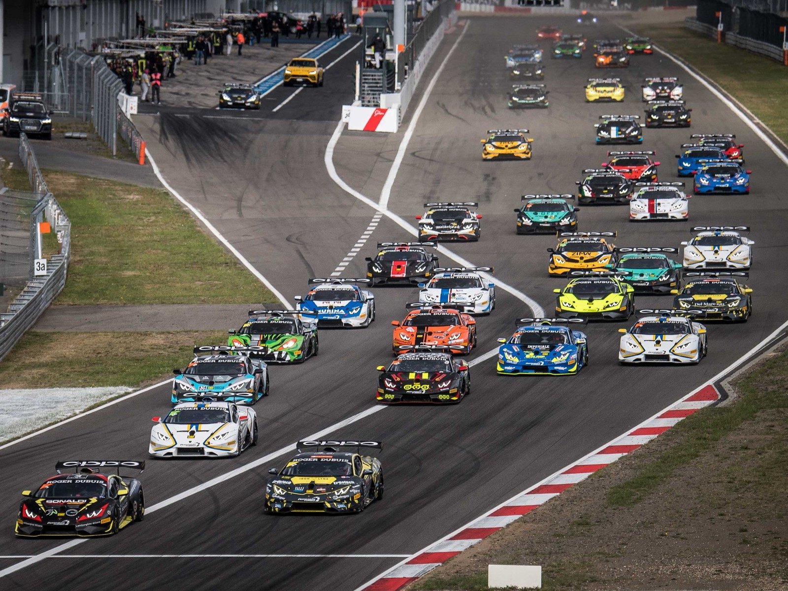 Full grid of Lamborghini Huracán Super Trofeo EVO race cars lined up on track with pit lane buildings in the background