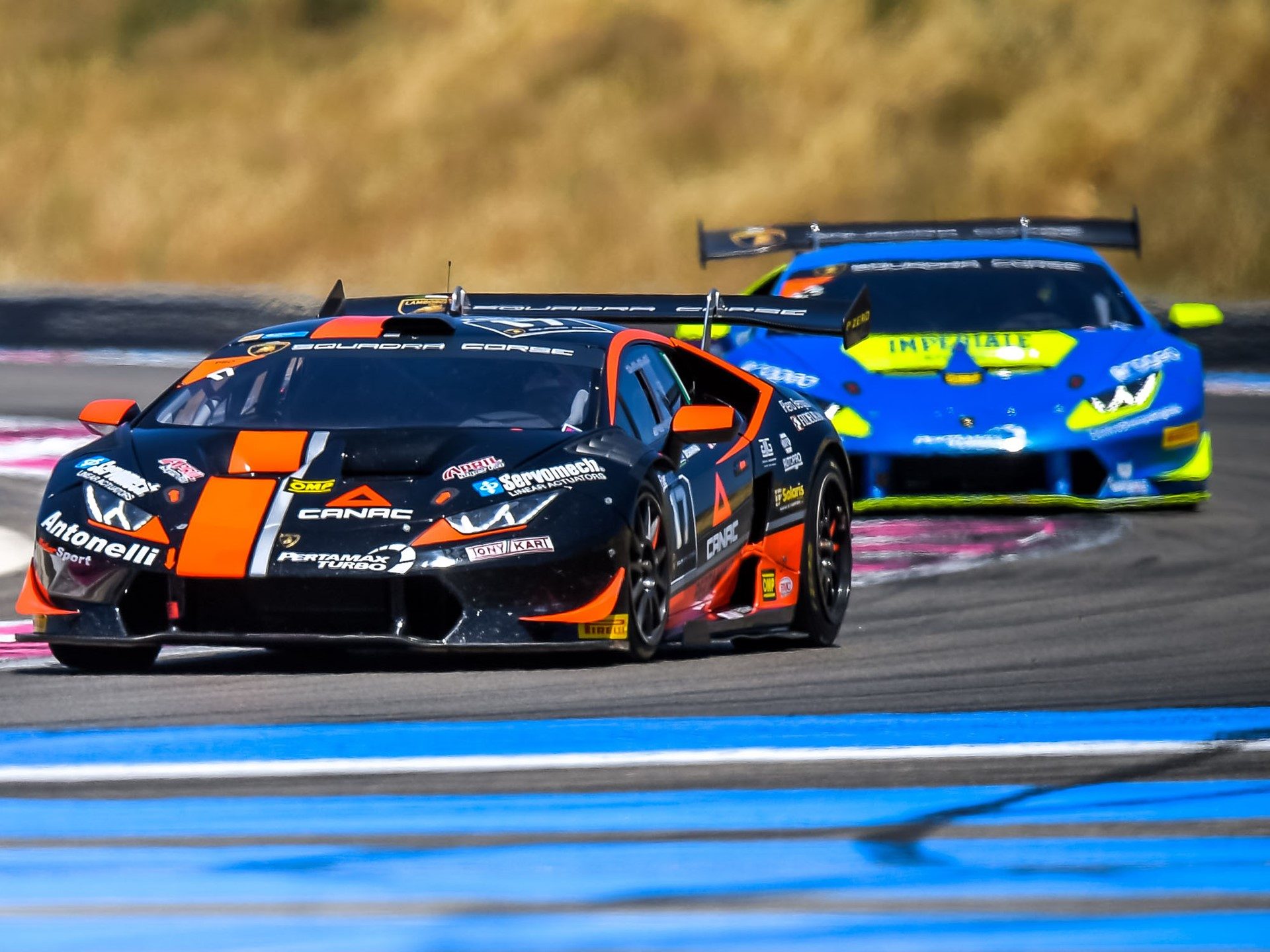 Two Lamborghini Huracán Super Trofeo race cars battling for position on a circuit, with the lead car in black and orange livery and the trailing car in blue and yellow