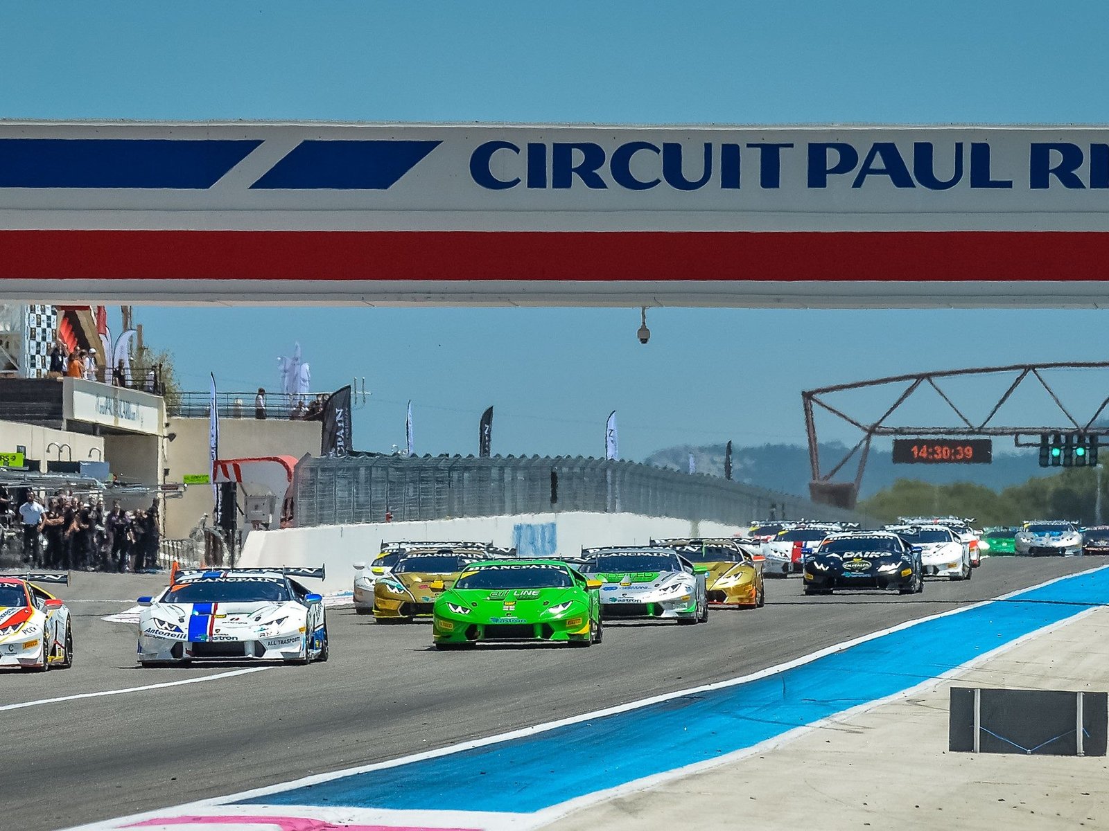 A fleet of Lamborghini Huracán Super Trofeo race cars in varied liveries lined up on the Circuit Paul Ricard racetrack under a clear blue sky