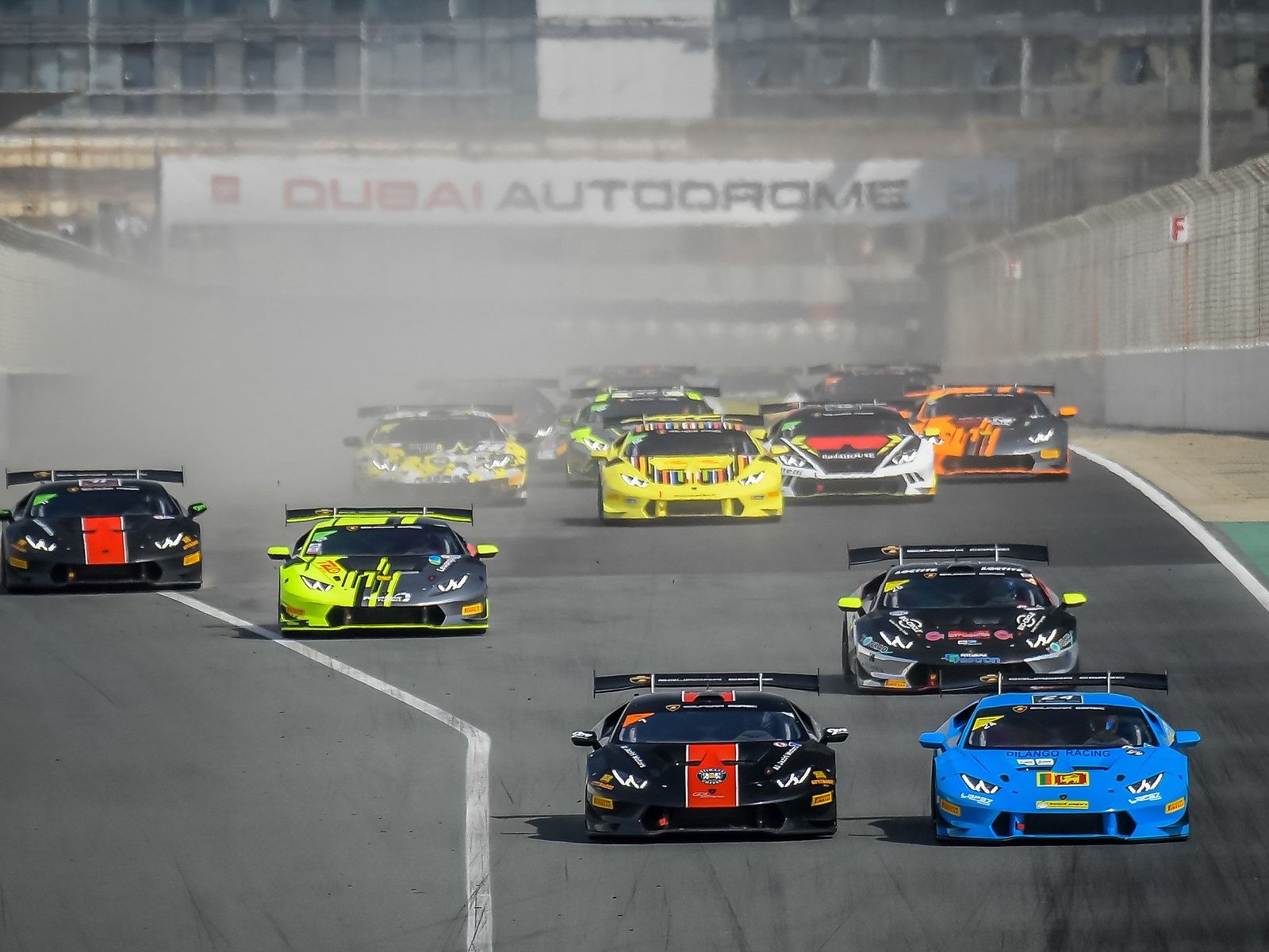 Multiple Lamborghini Huracán Super Trofeo race cars in black, yellow, and blue liveries competing on the Dubai Autodrome circuit