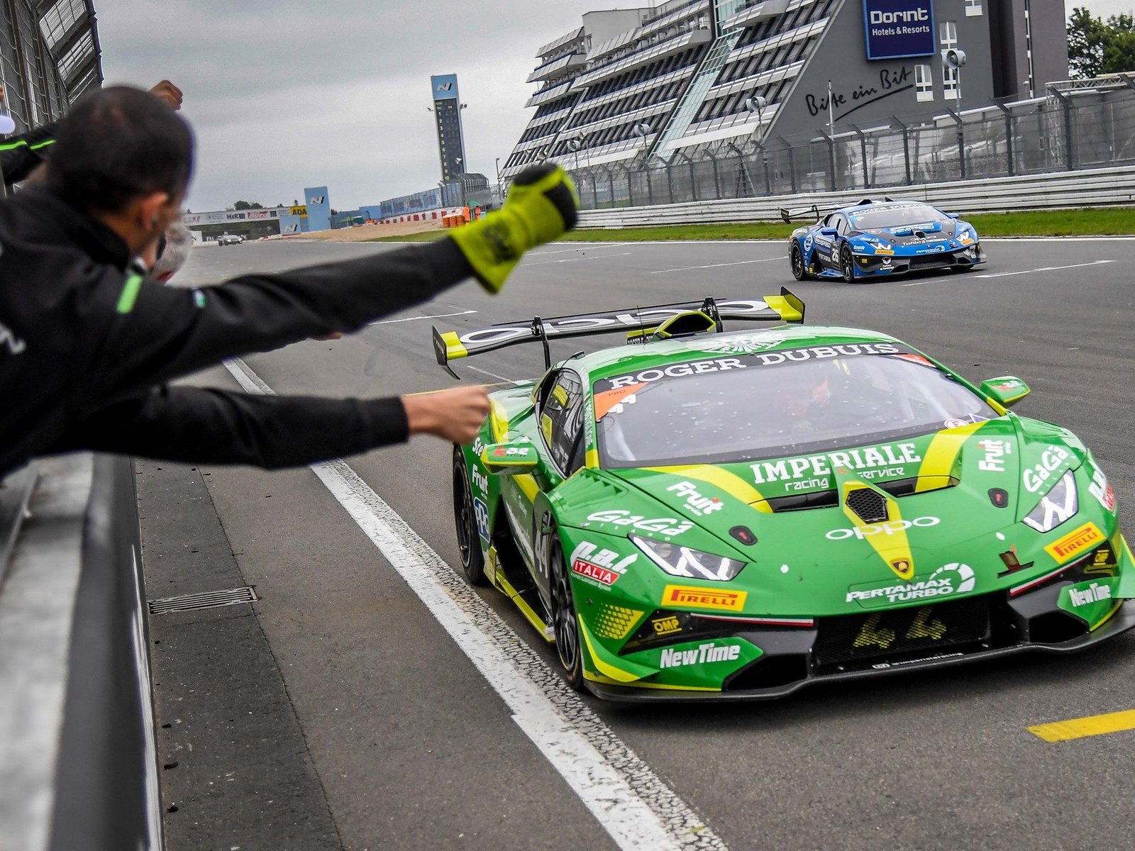 A pit crew member signals to a green and yellow Lamborghini Huracán Super Trofeo Evo race car on track at the Nürburgring, with a second Lamborghini visible in the background