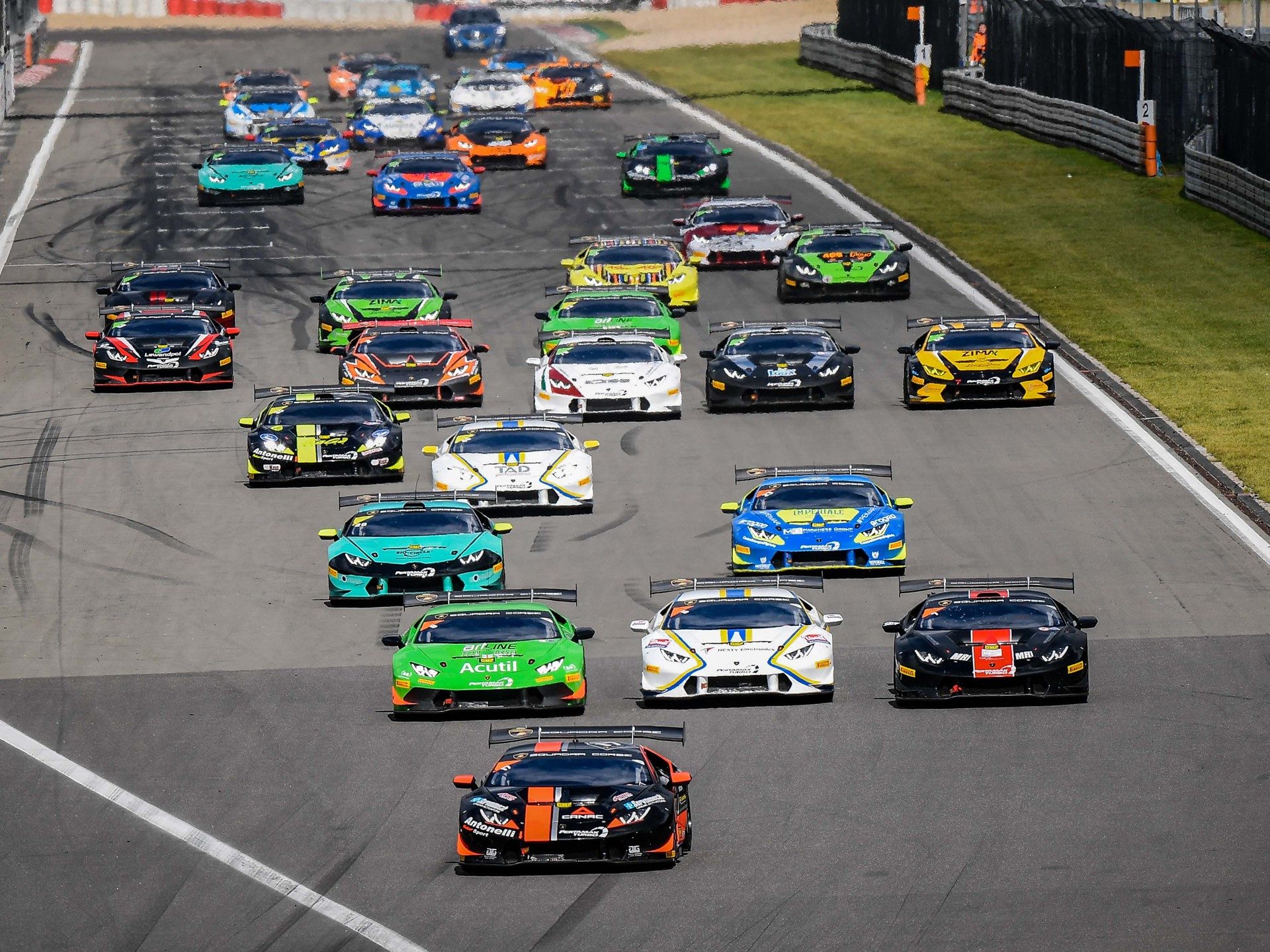 Numerous Lamborghini Huracán Super Trofeo race cars in vibrant liveries battle for position on the Nürburgring circuit under bright daylight