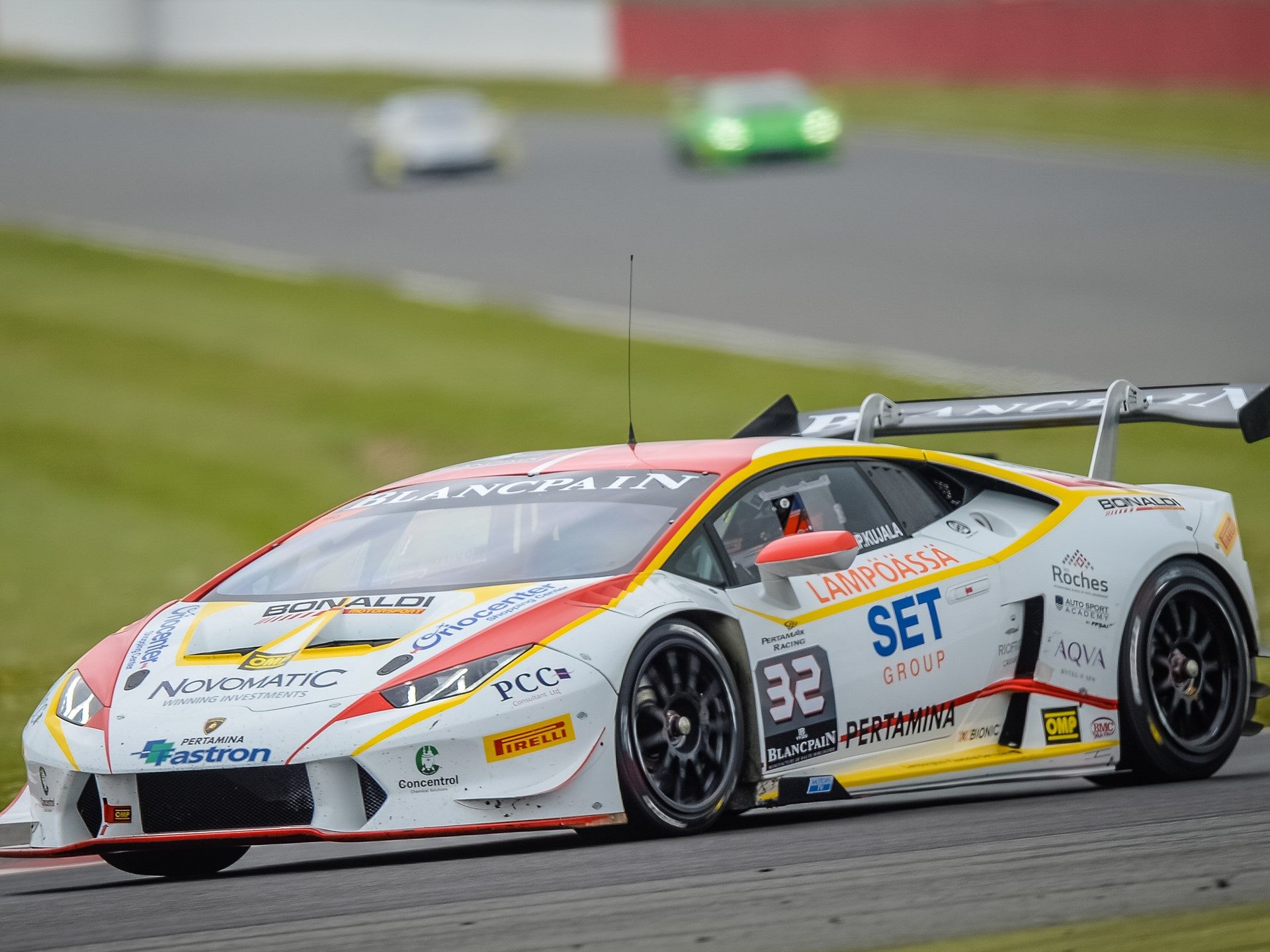 A white Lamborghini Huracán race car with red and yellow livery speeds through a corner on a racetrack, its large rear wing and aerodynamic body kit visible
