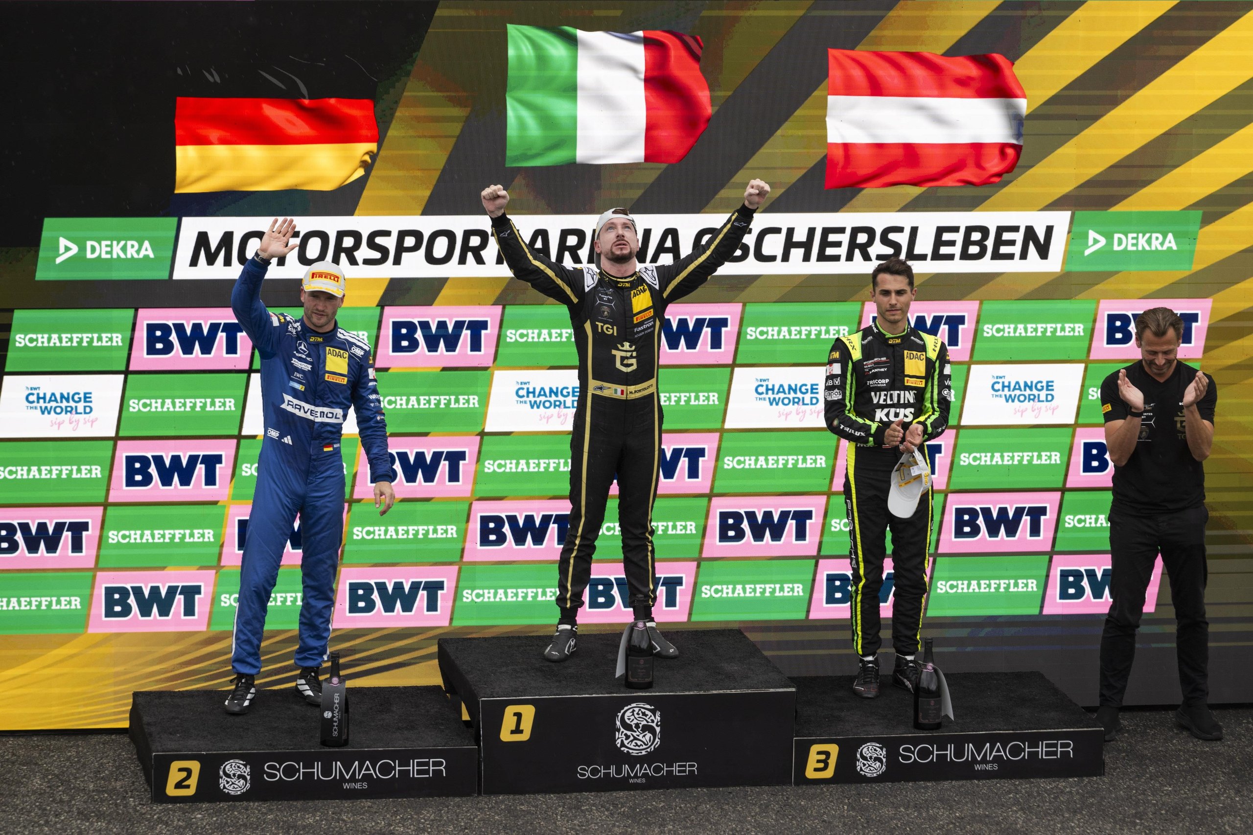 Three DTM race drivers on the podium celebrating with national flags of Italy, Germany, and Austria visible behind them