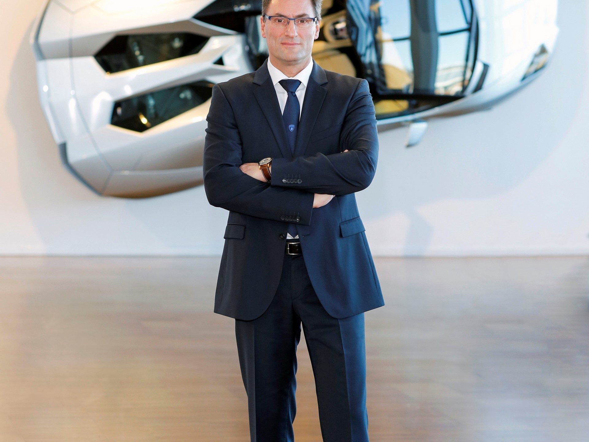 Werner Neuhold standing in a dark blue suit with arms crossed in front of a white angular Lamborghini design element
