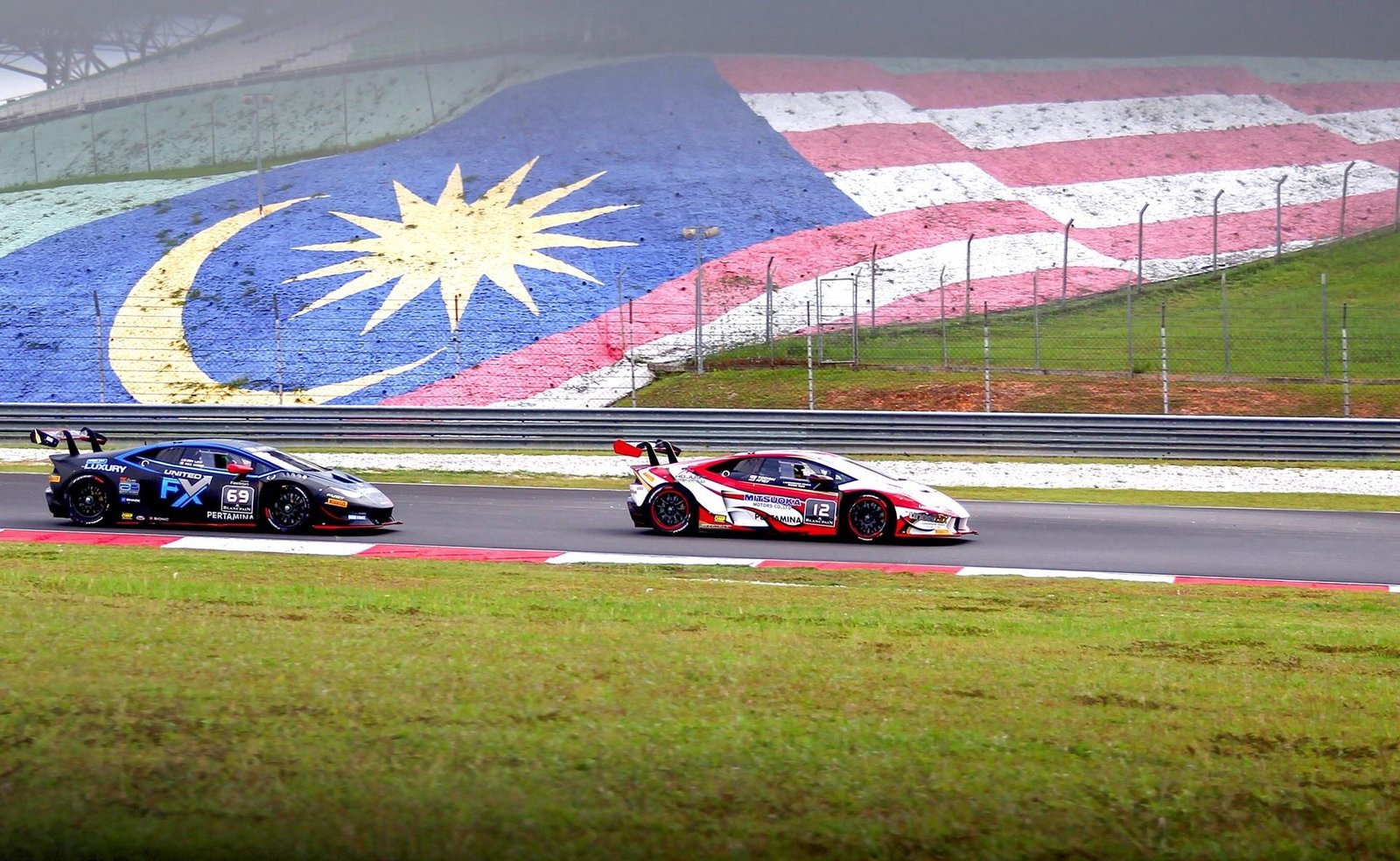 Two Lamborghini Huracán Super Trofeo race cars battle on track at Sepang with the Malaysian flag painted on a hillside behind them