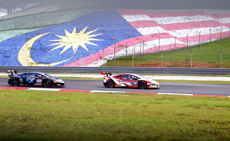 Two Lamborghini Huracán Super Trofeo race cars battle on track at Sepang with the Malaysian flag painted on a hillside behind them