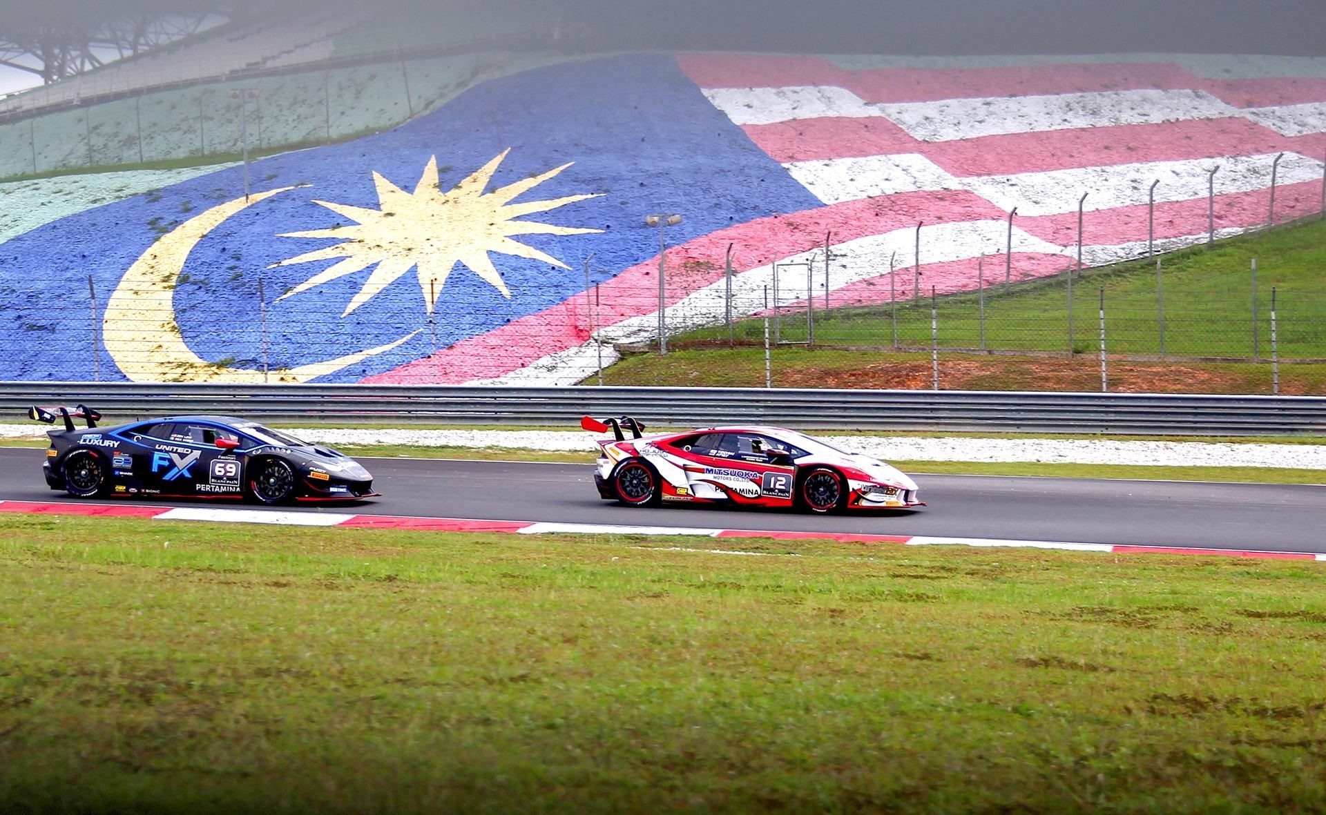 Two lamborghini huracán super trofeo race cars battle on track at sepang with the malaysian flag painted on a hillside behind them