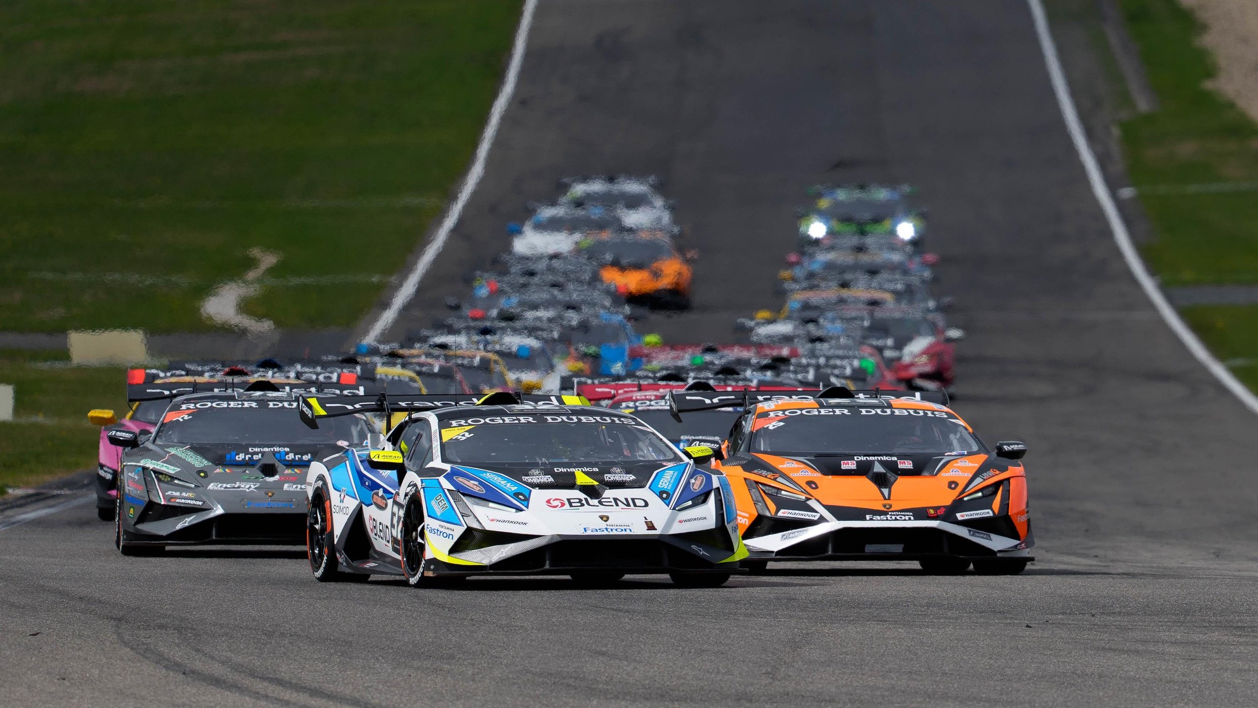 A pack of Lamborghini Huracán Super Trofeo EVO2 race cars in varied liveries racing down the Nürburgring straight with an orange car leading