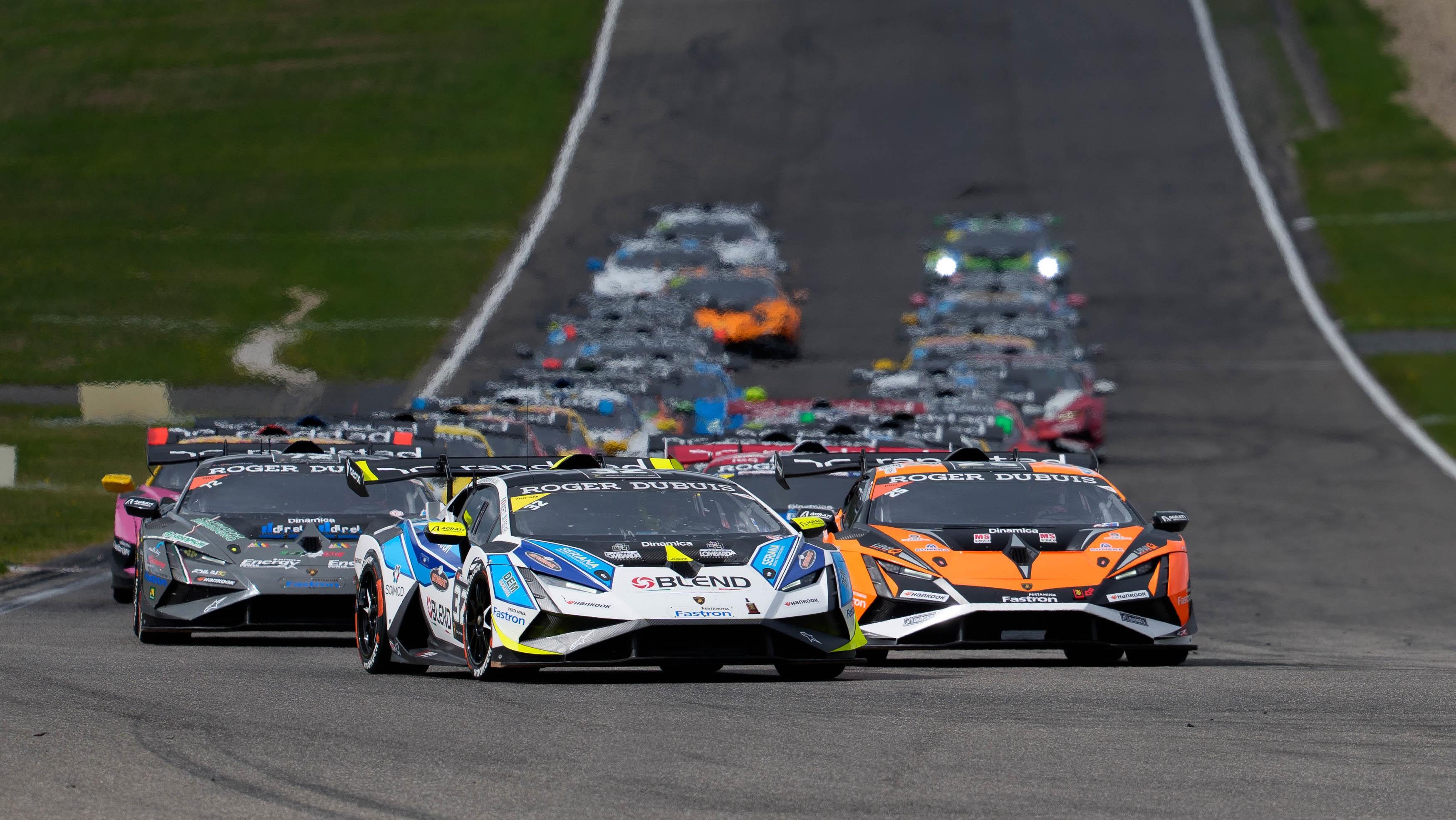 A pack of Lamborghini Huracán Super Trofeo EVO2 race cars in varied liveries racing down the Nürburgring straight with an orange car leading