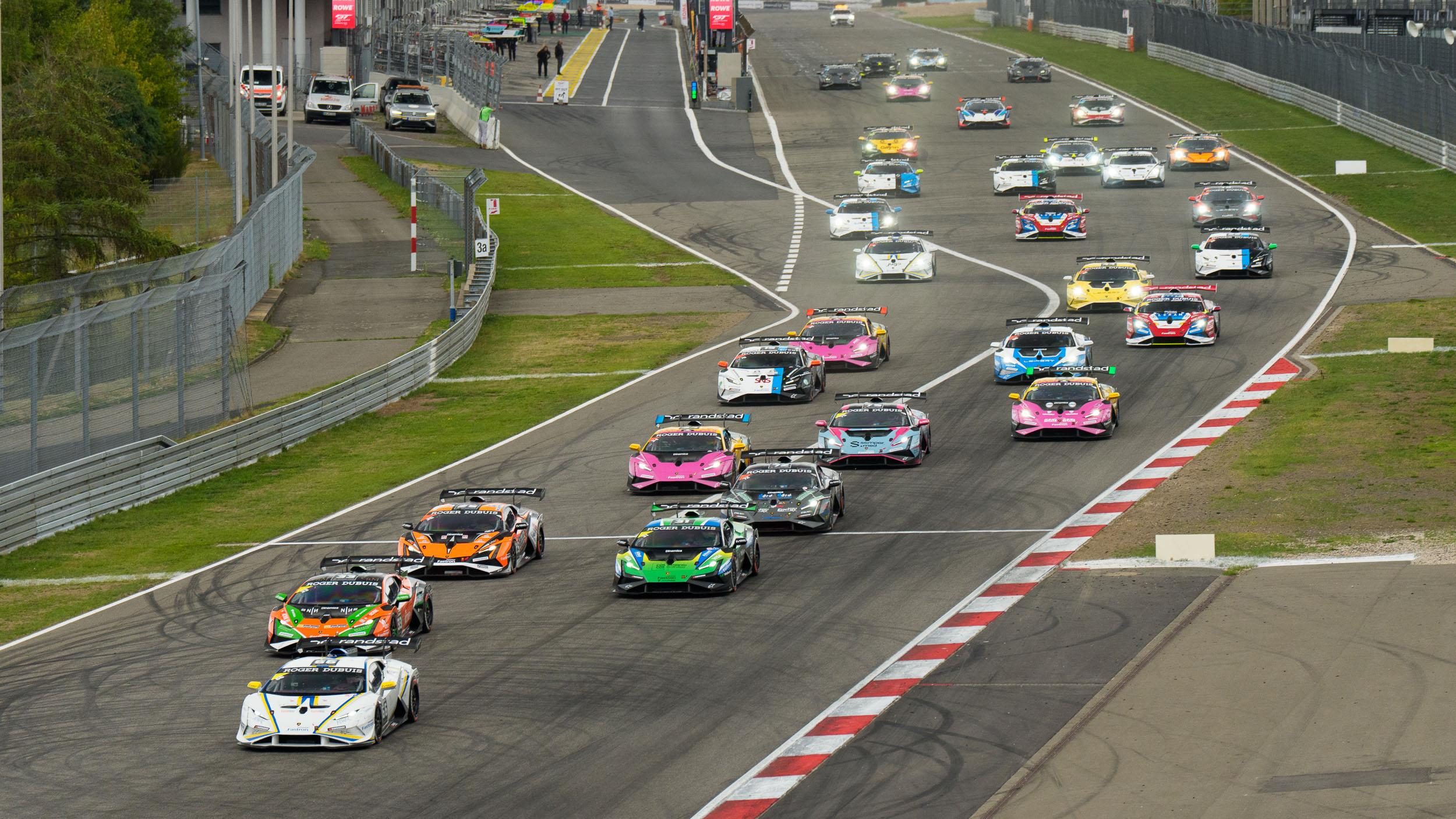 A large group of multi-colored Lamborghini Huracán Super Trofeo EVO2 race cars navigates a turn on a racetrack.