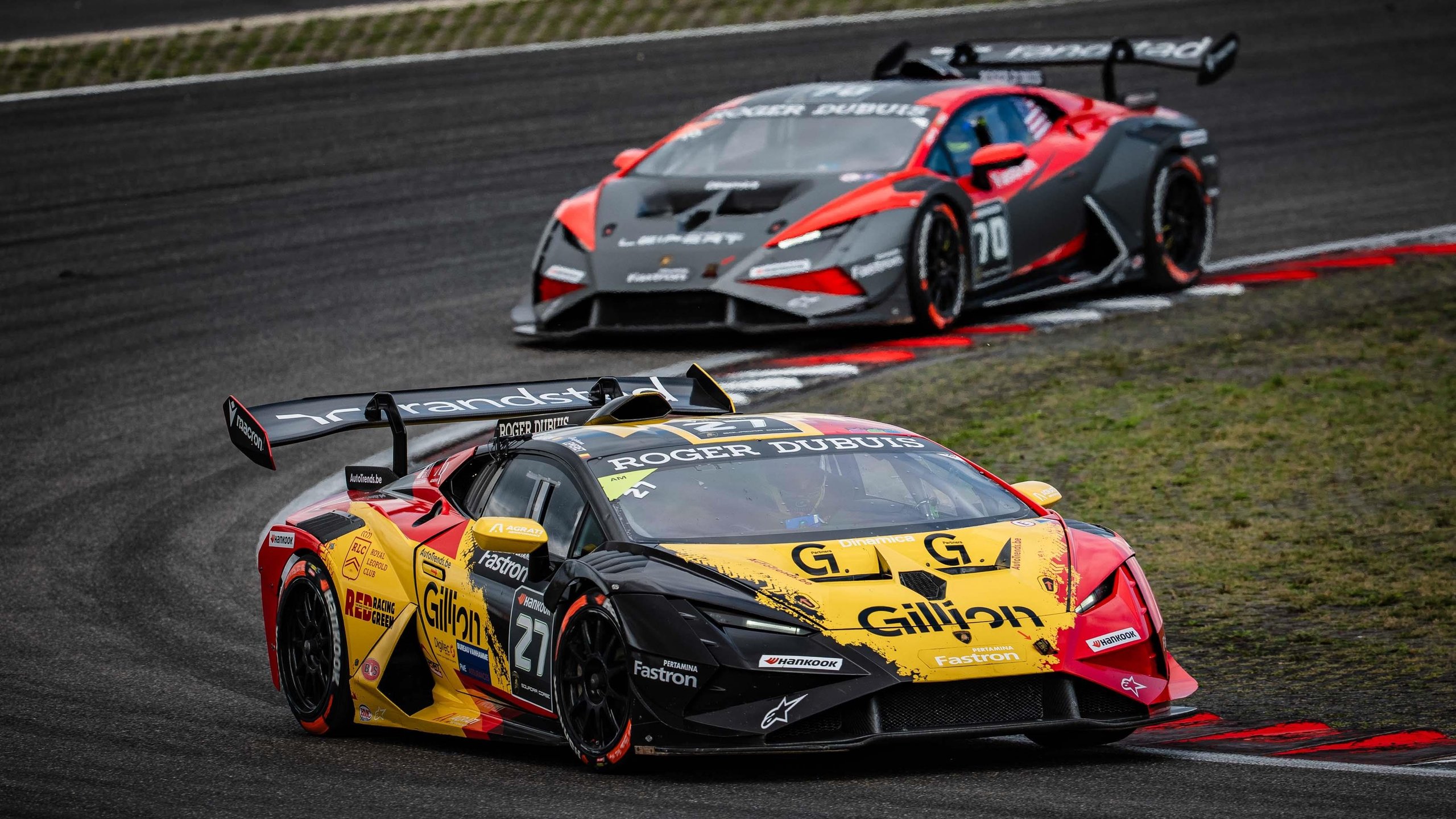 A multi-colored Lamborghini Huracán Super Trofeo EVO2 race car leads a grey and red Lamborghini Huracán Super Trofeo EVO2 on a racetrack.