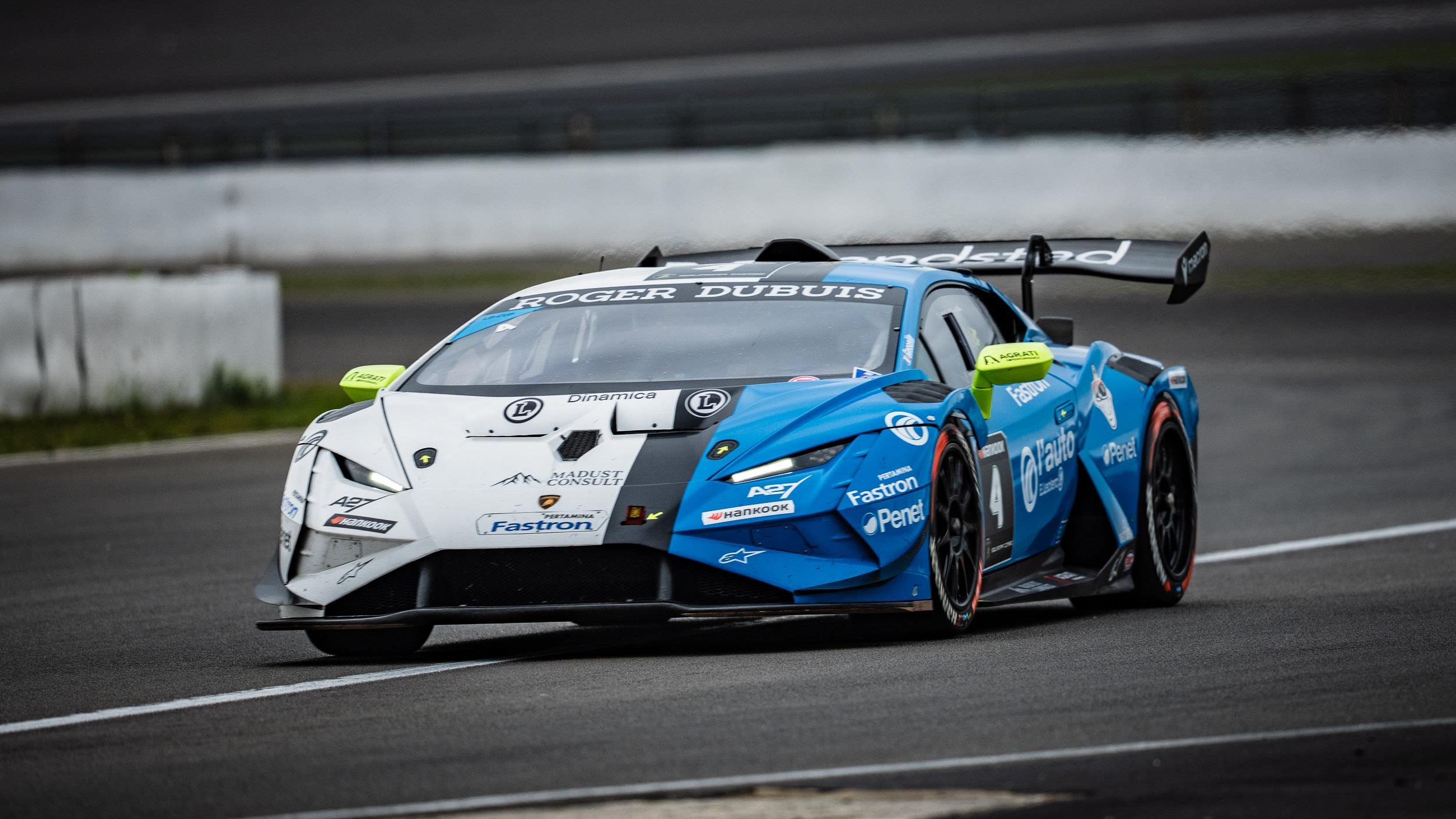 A blue, white, and black Lamborghini Huracán Super Trofeo EVO2 race car with green accents speeds on a racetrack under an overcast sky.