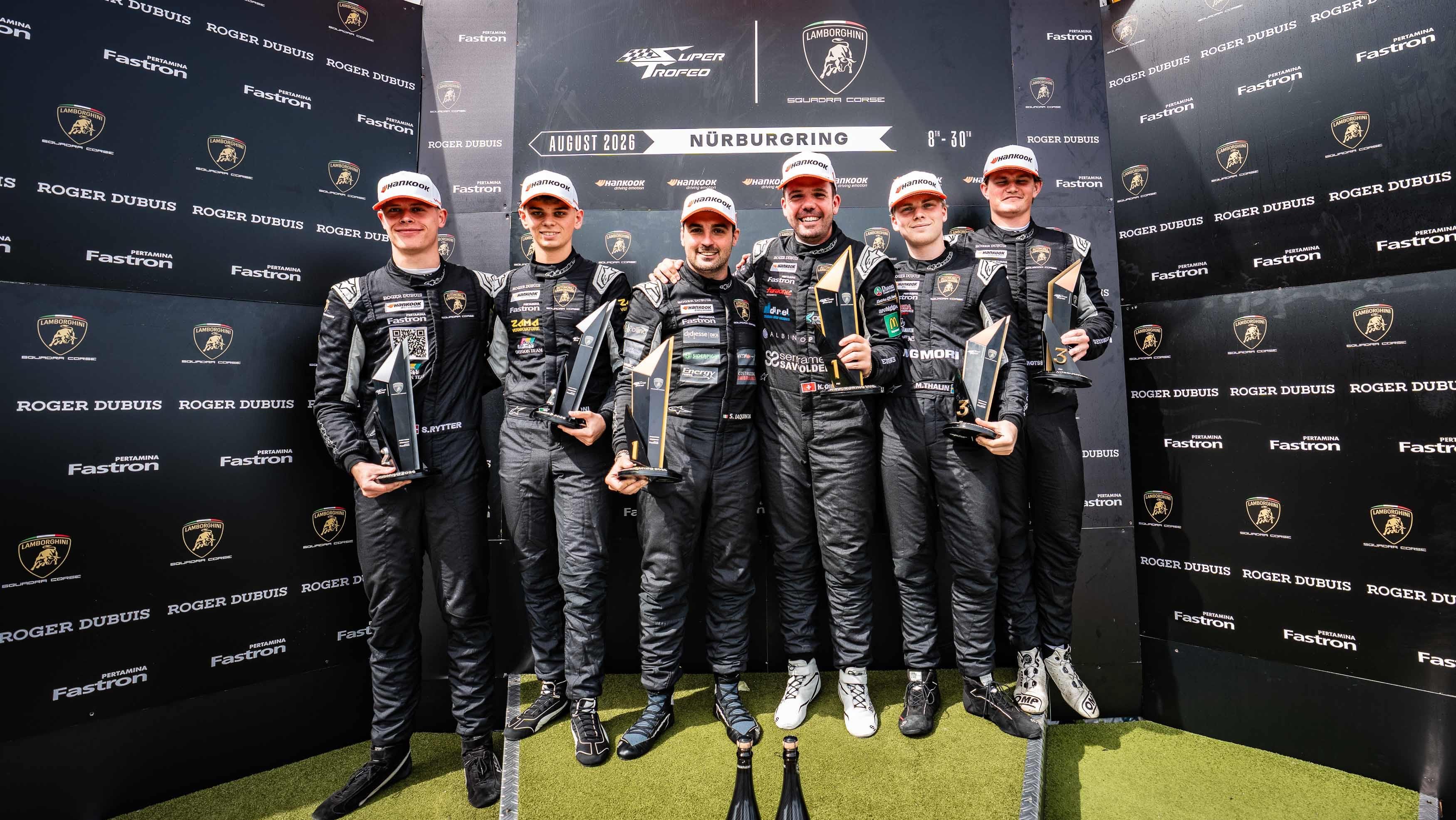 Six race car drivers in black racing suits celebrate on the Nürburgring podium with trophies
