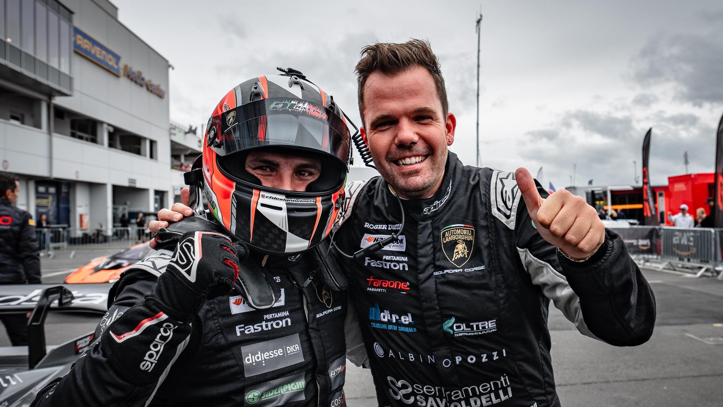 Two men, one in a racing helmet and the other giving a thumbs-up, pose for a photo at a racetrack event, both wearing black racing attire with sponsor logos.