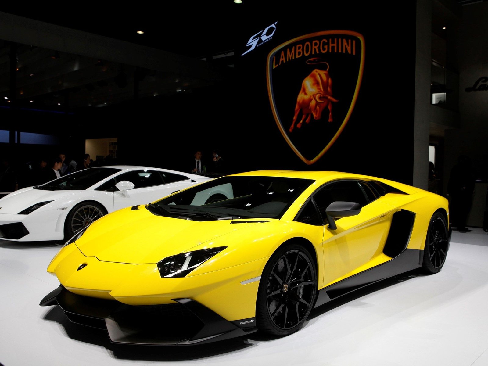Bright yellow Lamborghini Aventador LP 720-4 50 Anniversario displayed at an auto show with a white Gallardo visible in the background and 50 Anniversario branding on the backdrop