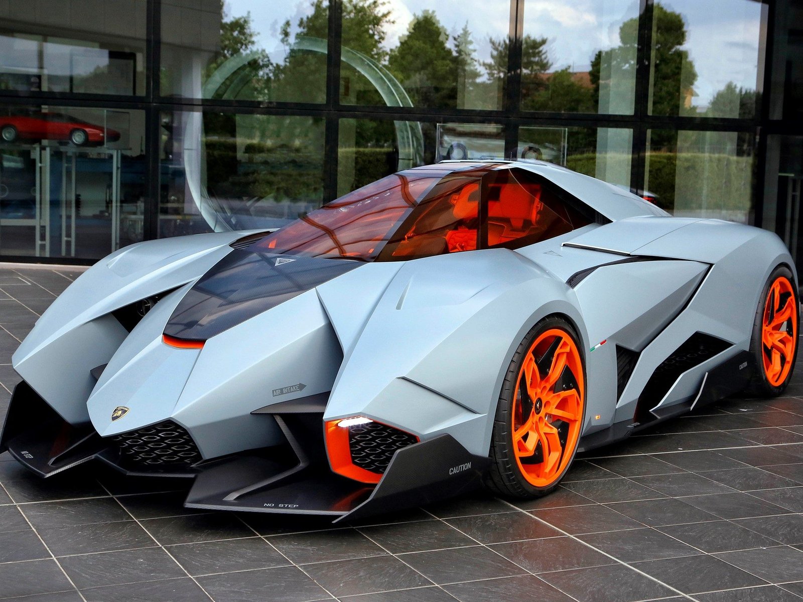 The Lamborghini Egoista concept car in matte grey with orange wheel accents and cockpit tinting, parked outside a modern glass building at Lamborghini headquarters