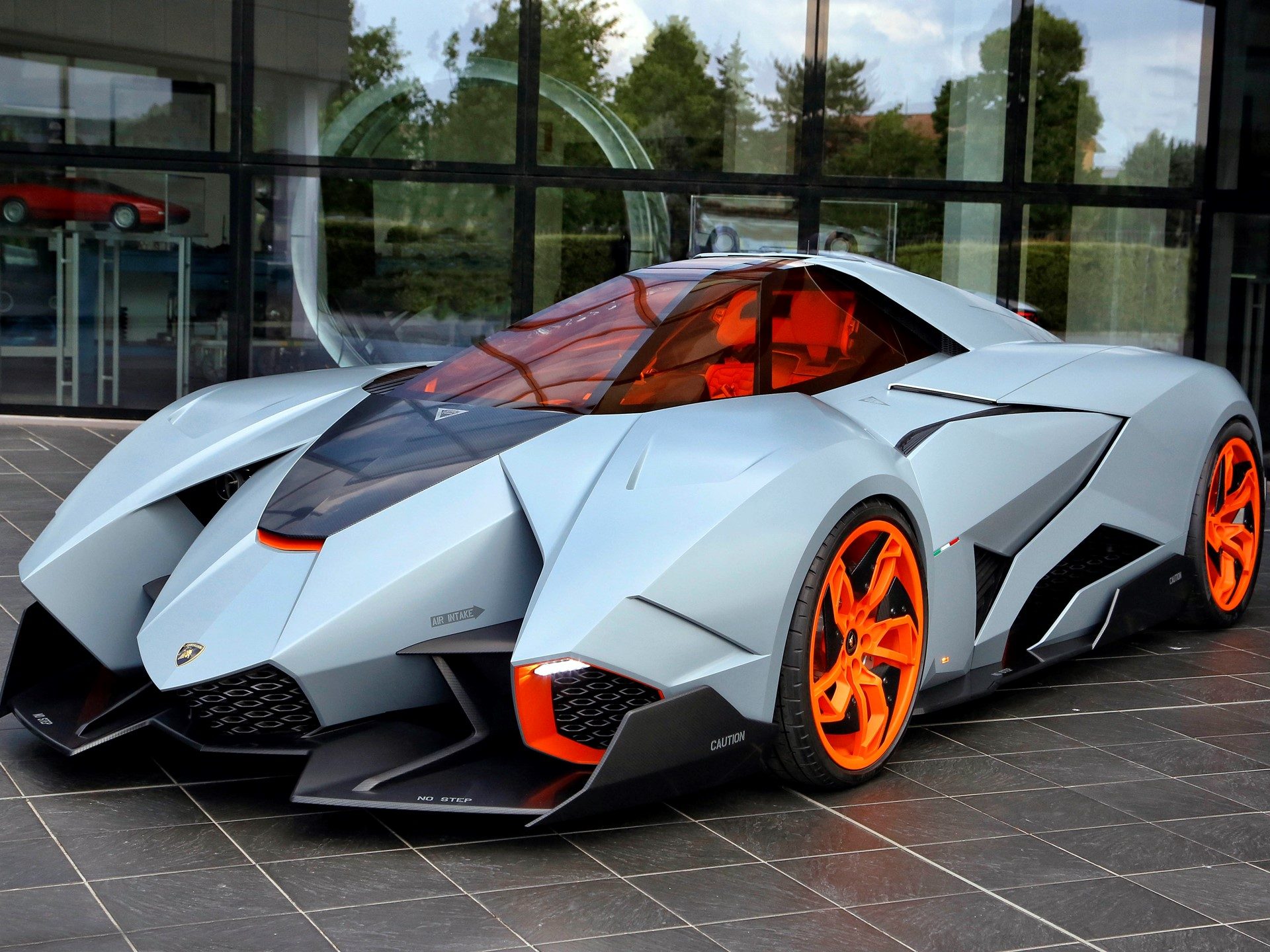 The Lamborghini Egoista concept car in matte grey with orange wheel accents and cockpit tinting, parked outside a modern glass building at Lamborghini headquarters