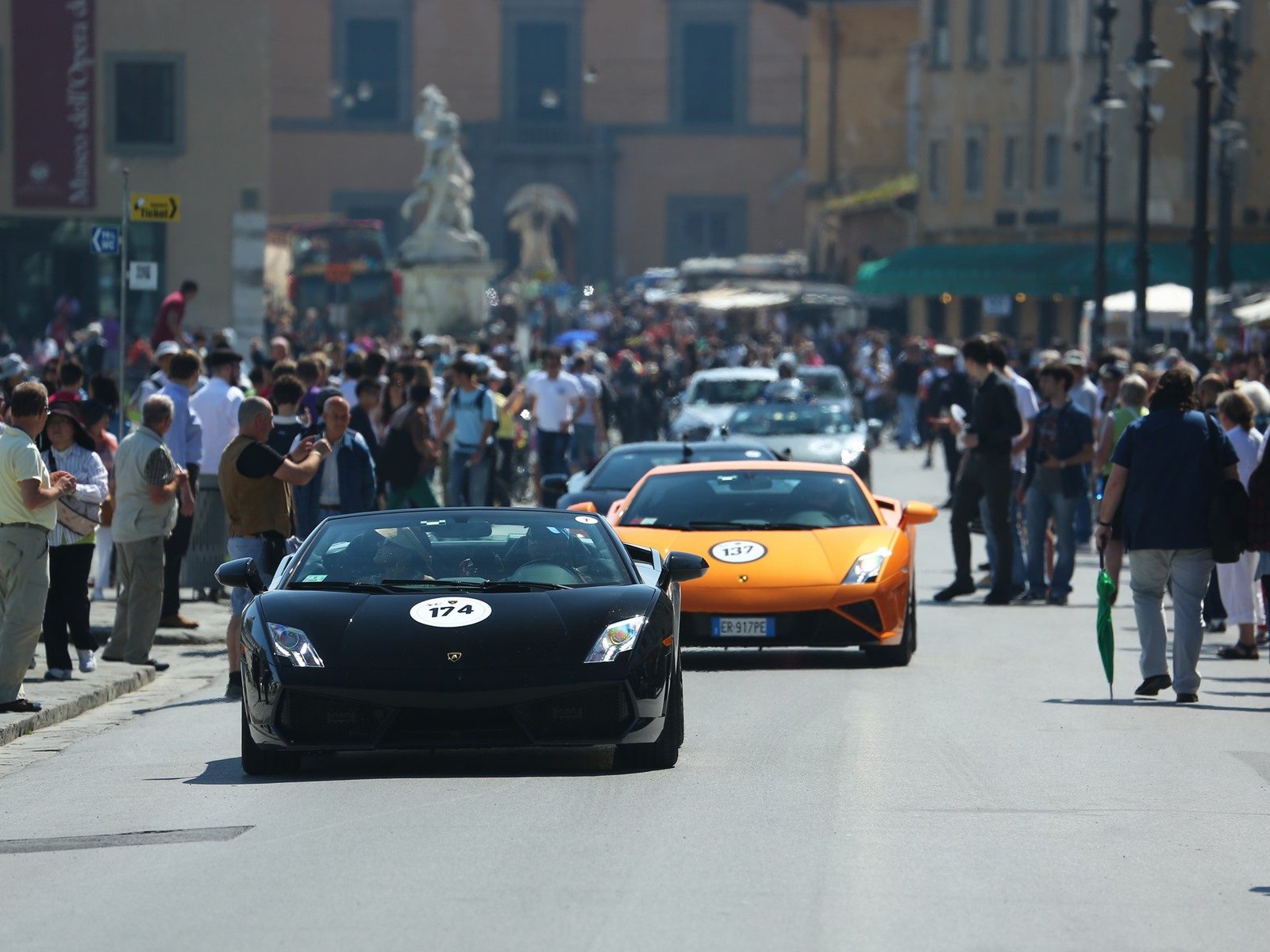 A black Lamborghini Gallardo leads an orange Gallardo through a spectator-lined street during the 50th Anniversary Grand Tour
