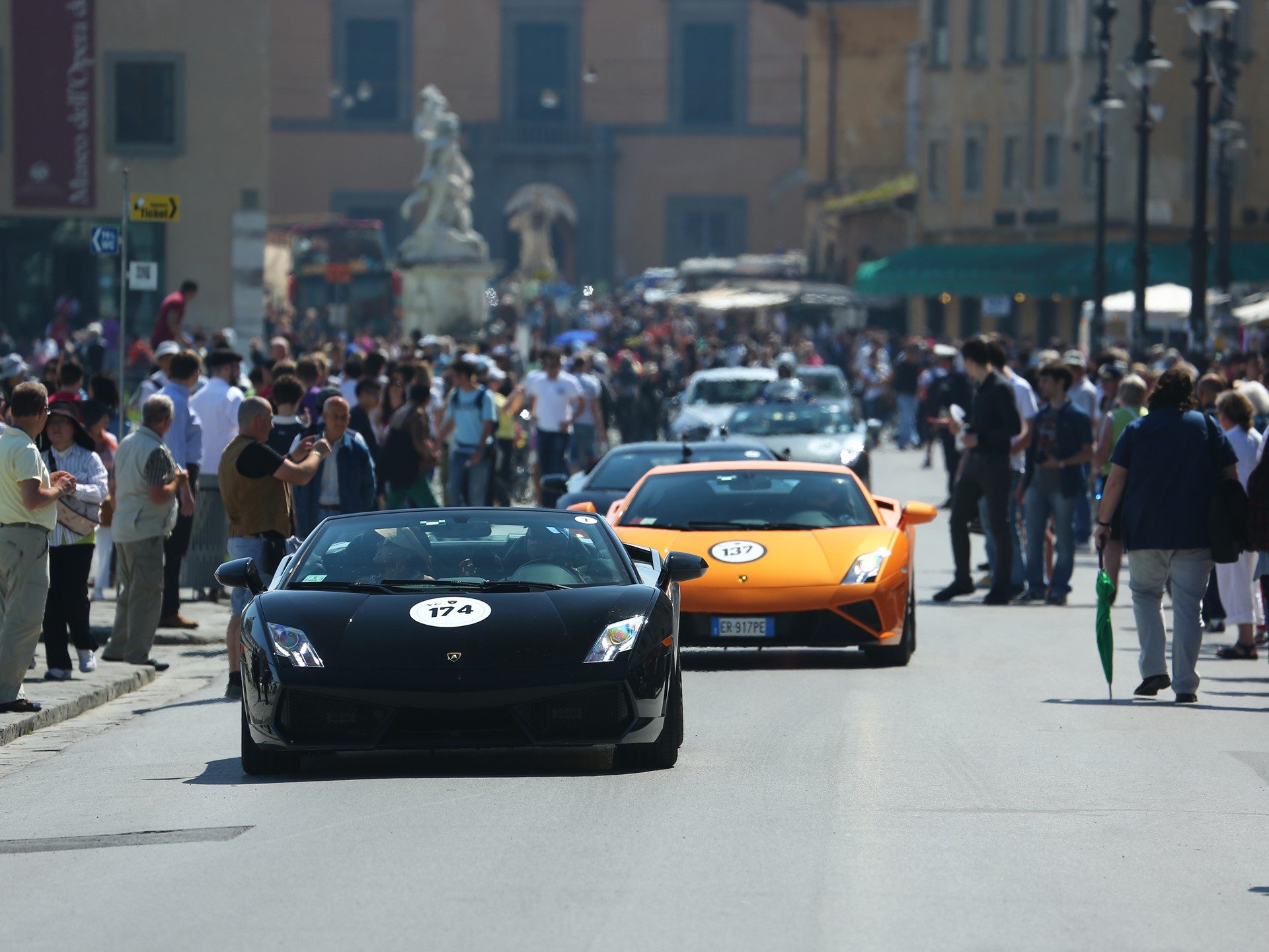 A black Lamborghini Gallardo leads an orange Gallardo through a spectator-lined street during the 50th Anniversary Grand Tour