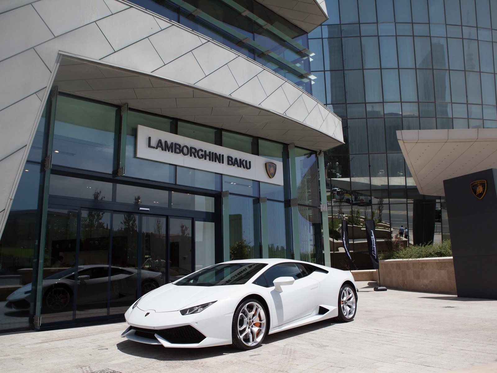 A white Lamborghini Huracán LP 610-4 parked in front of the glass-facade Lamborghini Baku dealership with clear blue sky overhead