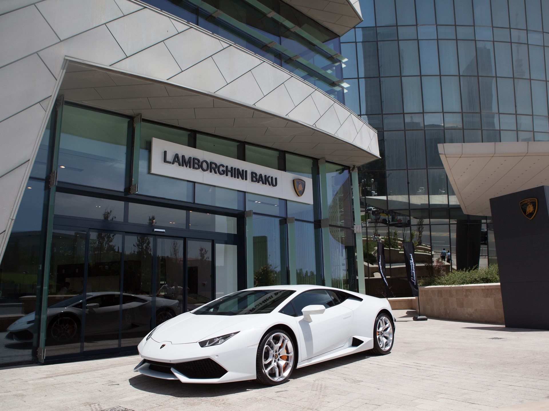 A white Lamborghini Huracán LP 610-4 parked in front of the glass-facade Lamborghini Baku dealership with clear blue sky overhead