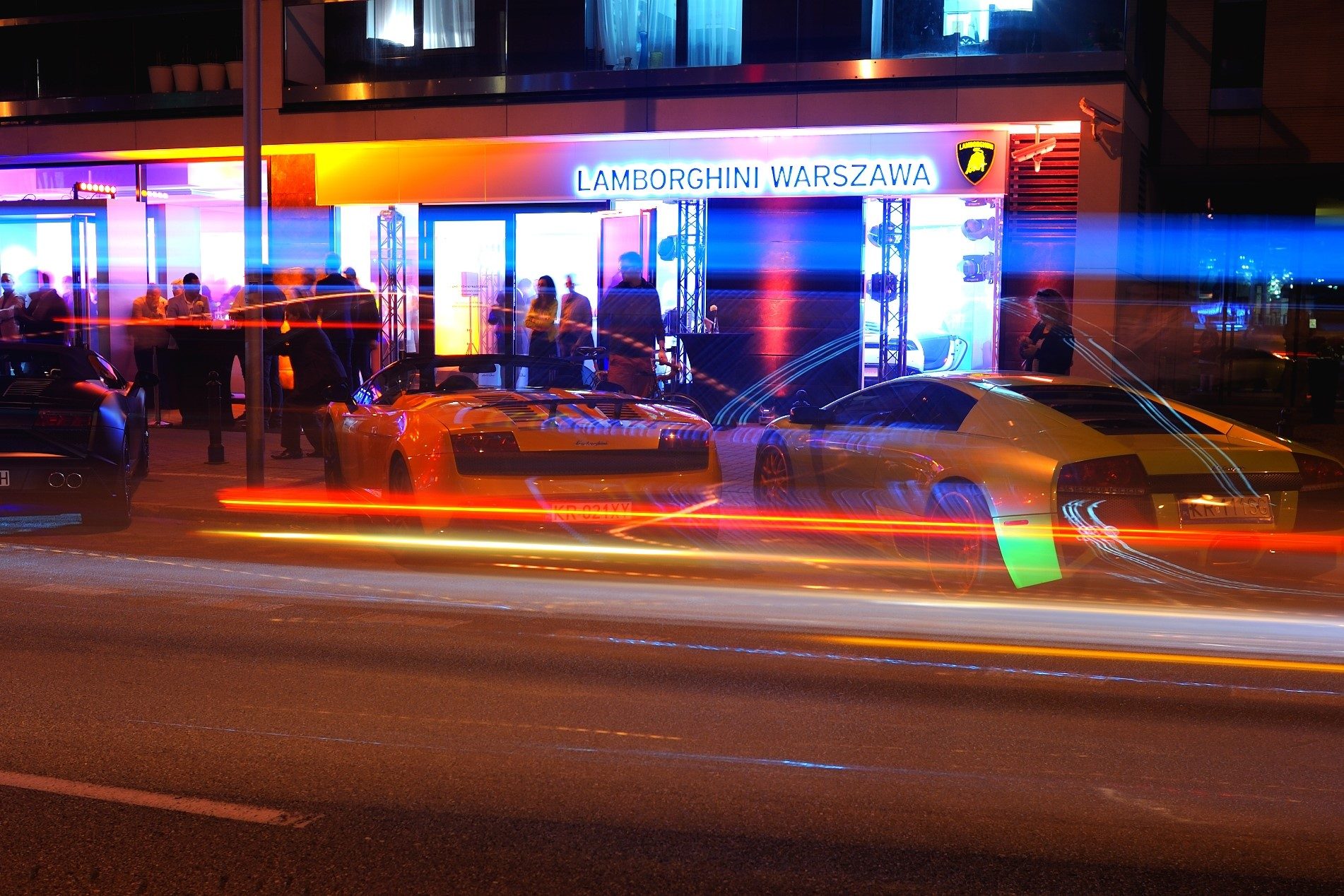 Orange Gallardo and yellow Murciélago outside the Lamborghini Warsaw showroom at night with visible event activity inside