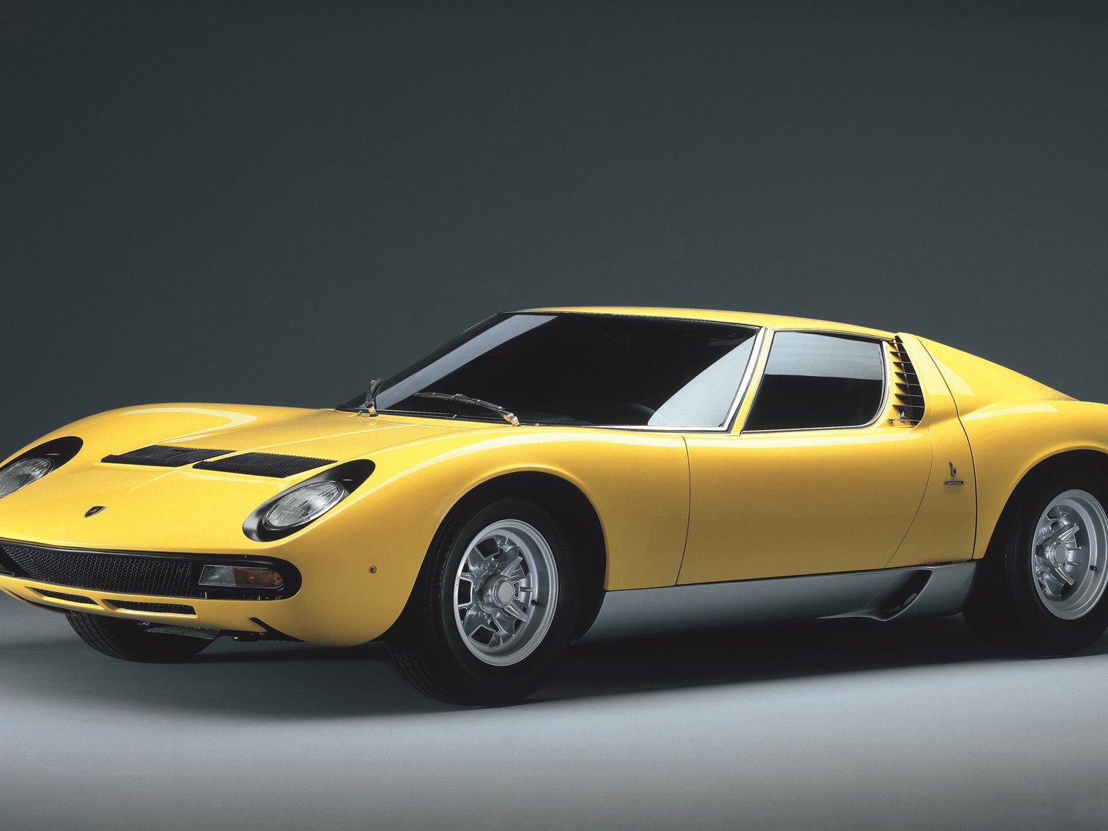 Classic yellow Lamborghini Miura in studio setting showing sleek mid-engine profile, pop-up headlights, and louvered rear window