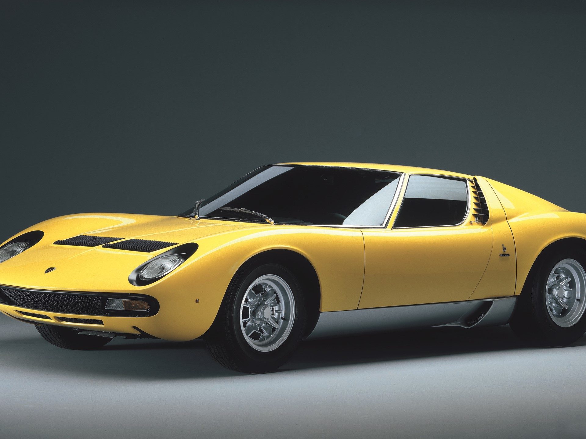 Classic yellow Lamborghini Miura in studio setting showing sleek mid-engine profile, pop-up headlights, and louvered rear window