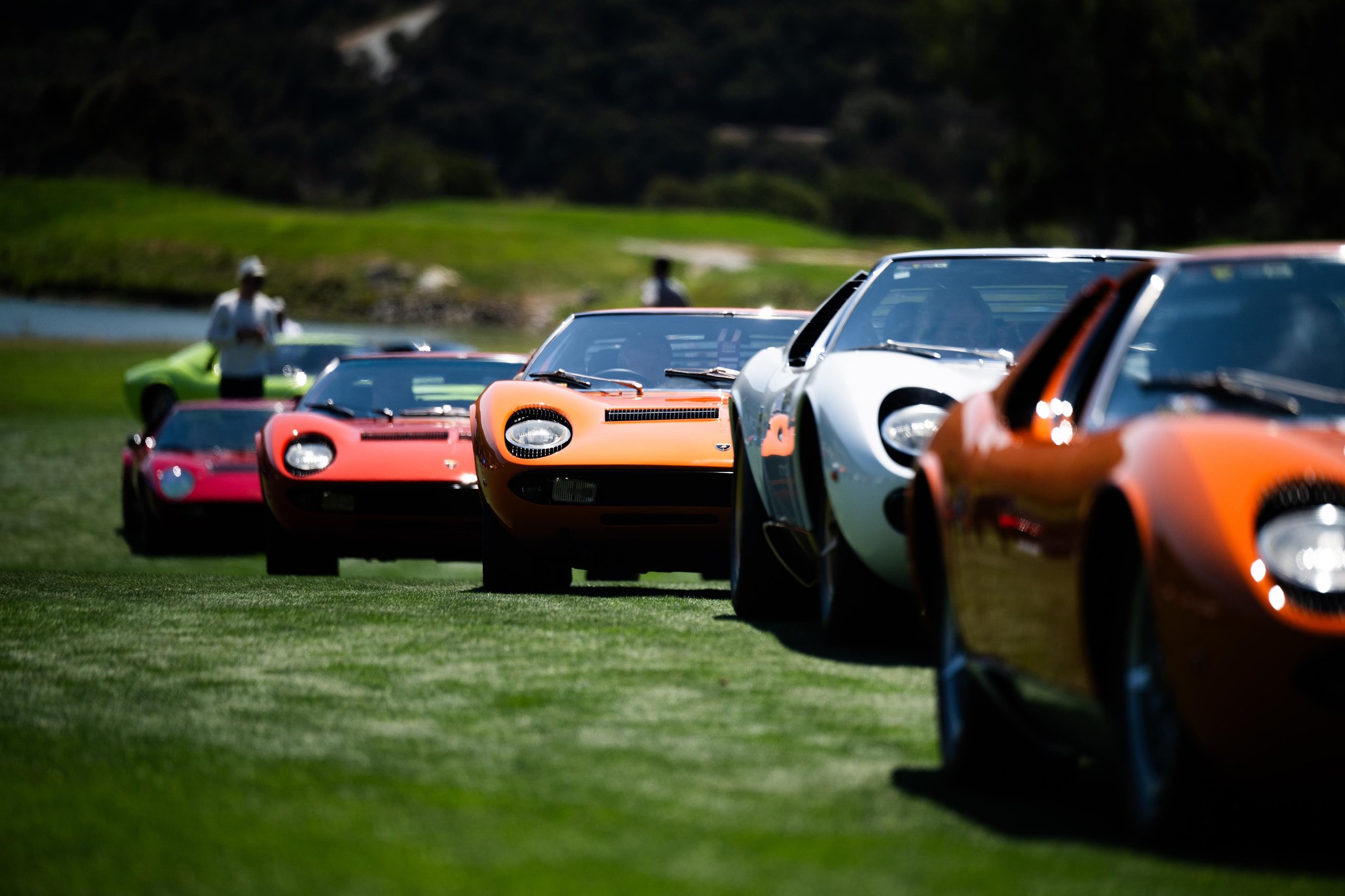 A convoy of classic Lamborghini Miuras in orange, white, red, and green driving across a green field at Monterey Car Week