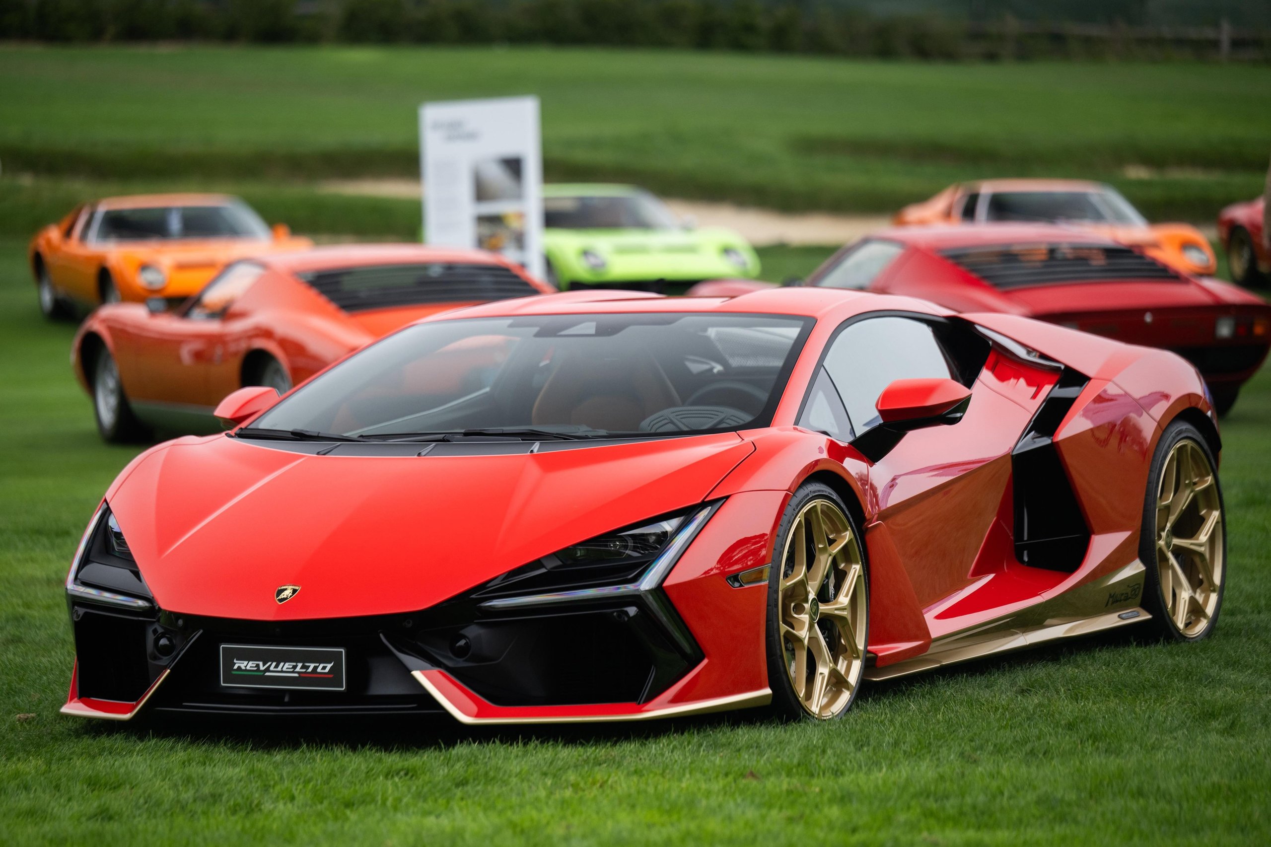 Red Lamborghini Revuelto with gold wheels displayed on a lawn with classic Miuras visible in the blurred background