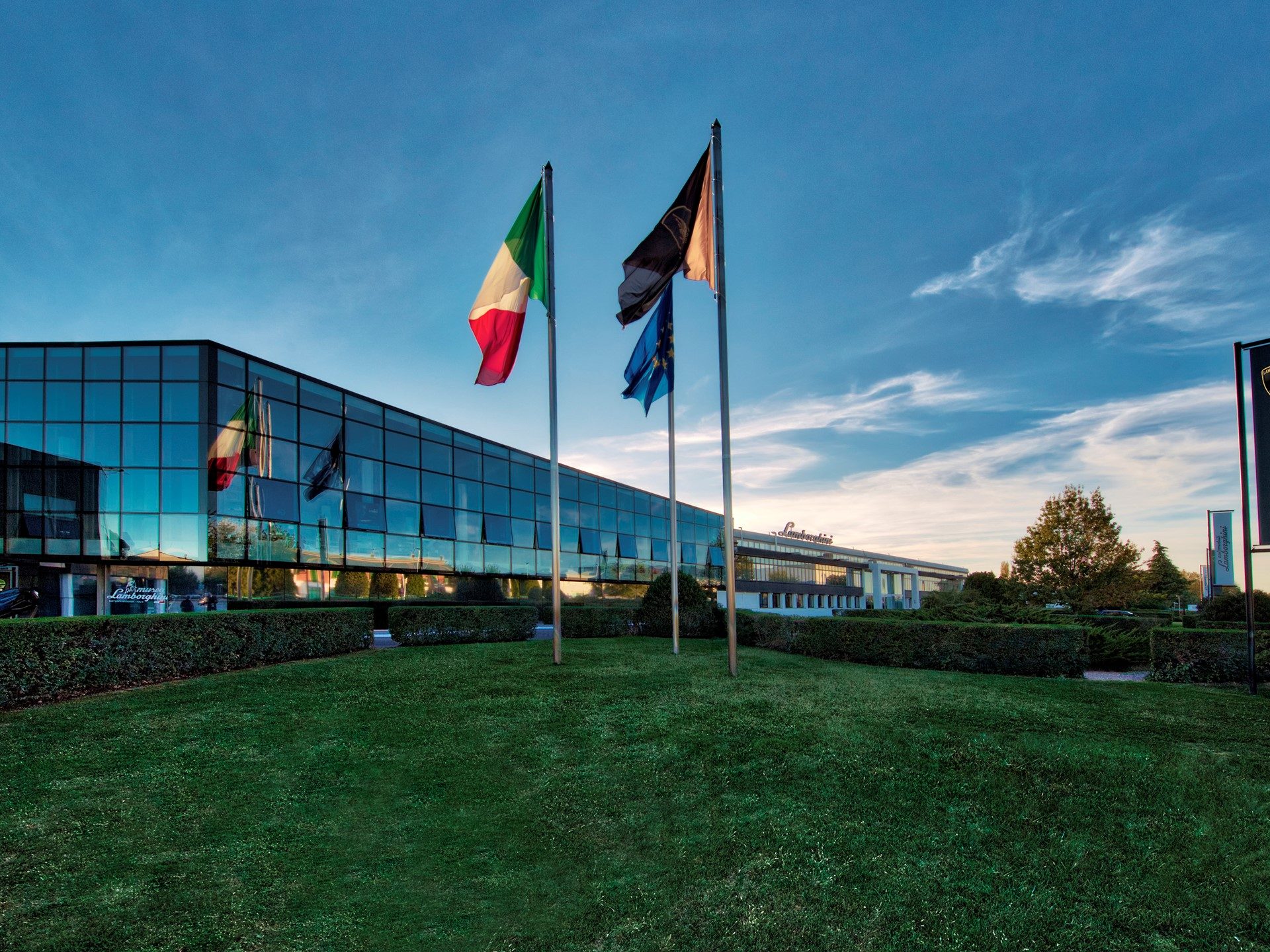 Lamborghini factory headquarters in Sant'Agata Bolognese with Italian, EU, and Lamborghini flags flying in front of the modern glass facade