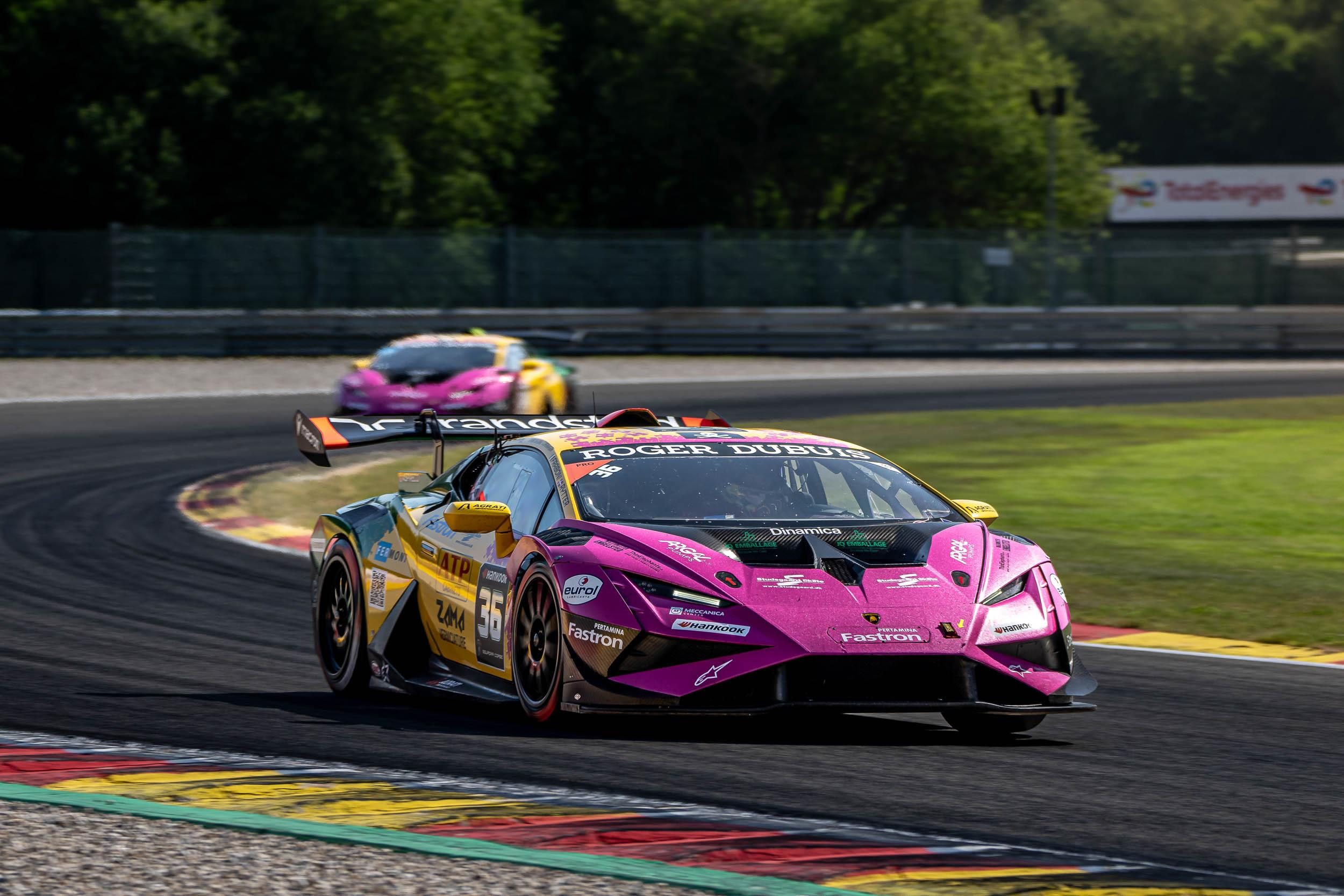 A Lamborghini Huracán Super Trofeo EVO2 race car in pink, yellow, and green livery leads another car around a sweeping turn